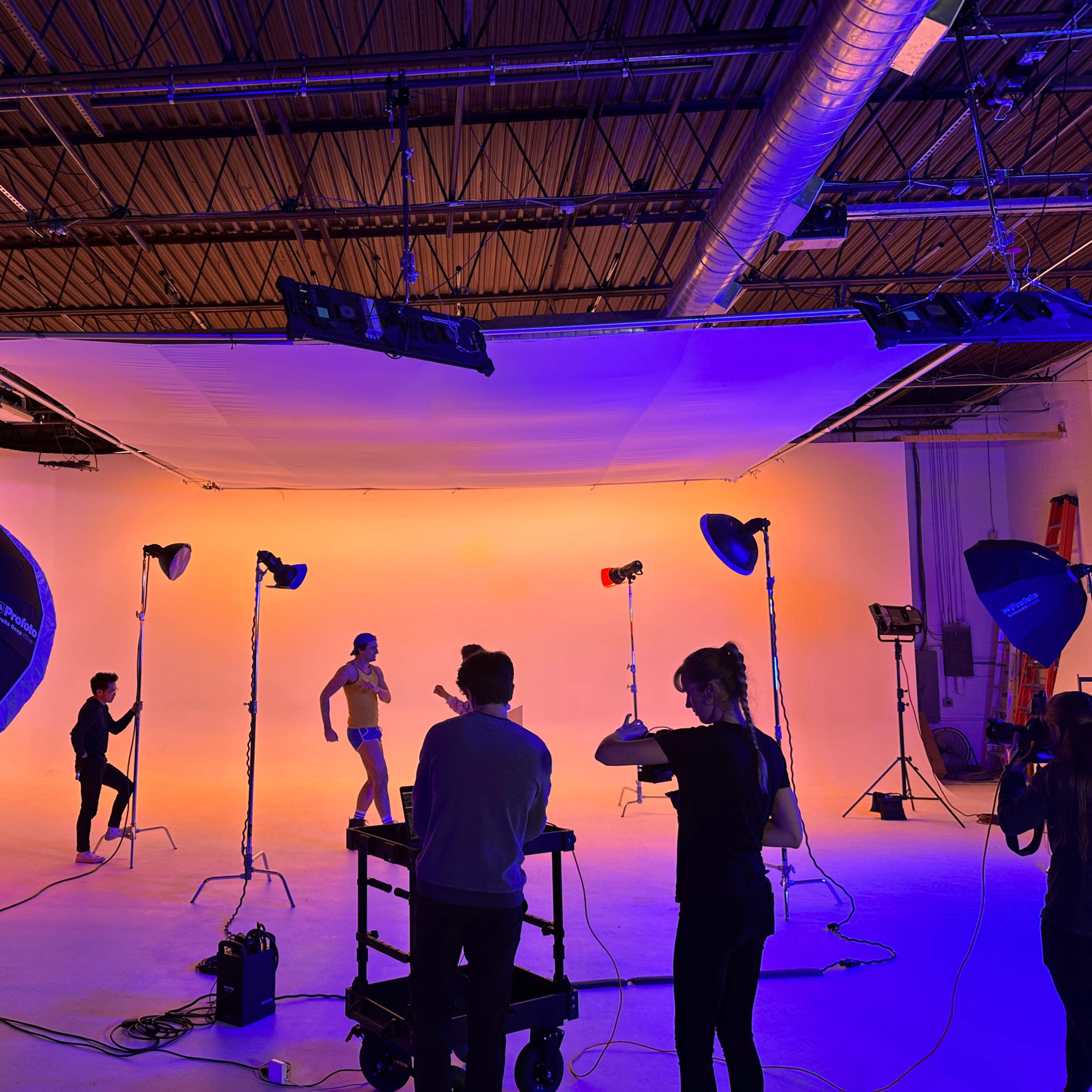 A studio space is set up for a photoshoot, featuring lighting equipment, a backdrop, and several individuals posing and adjusting settings.