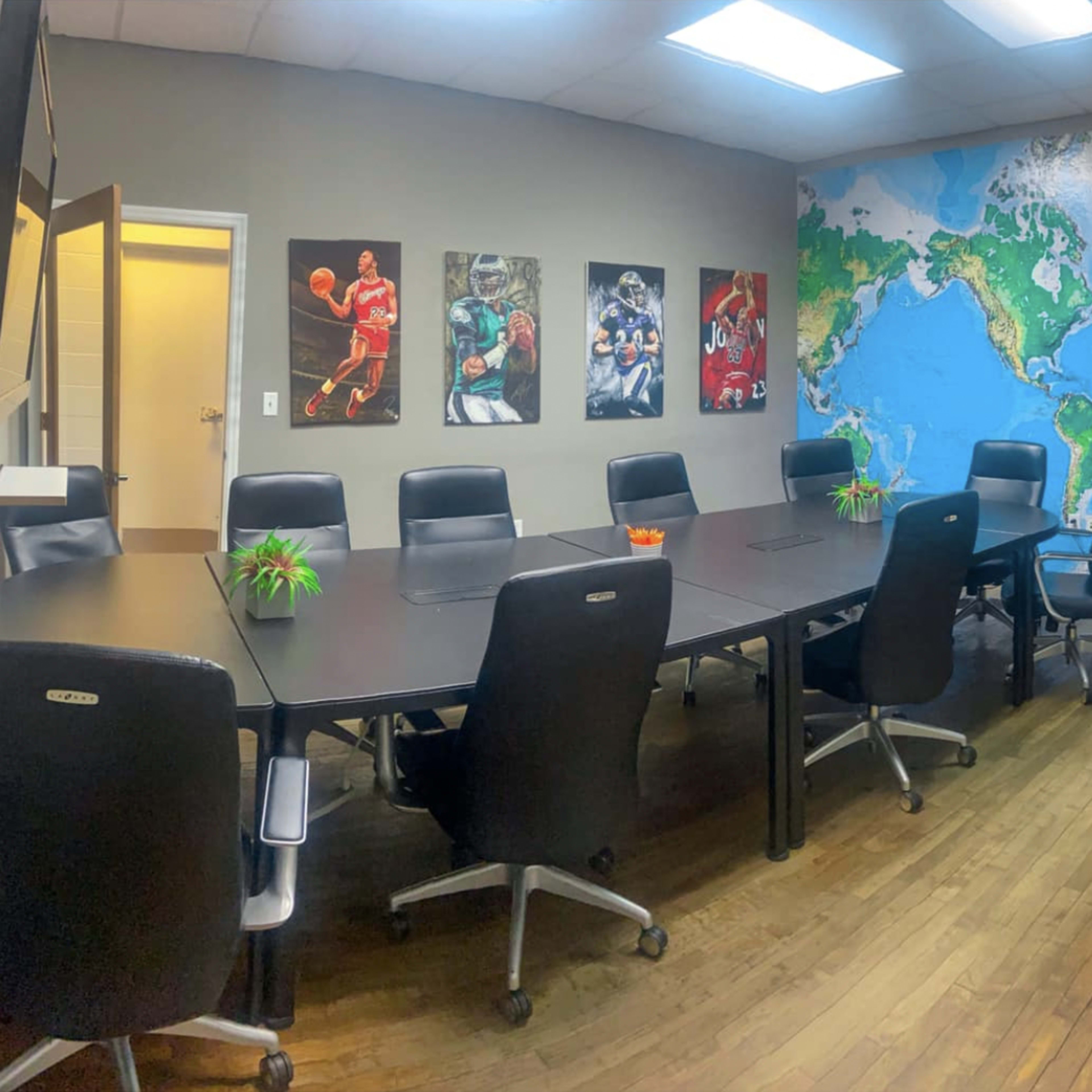 A conference room is furnished with a large table surrounded by black chairs, featuring a world map mural on one wall and sports-themed artwork on another.