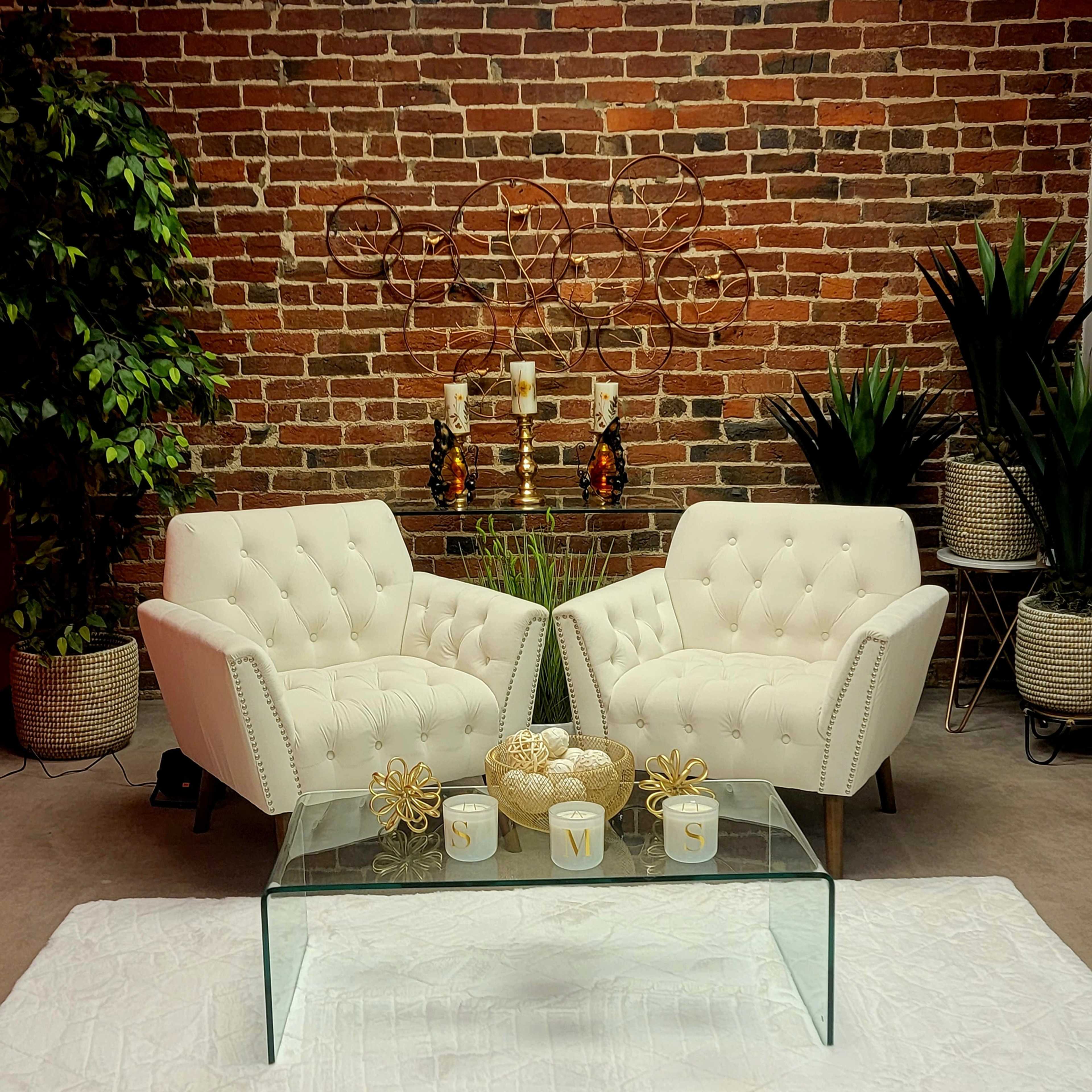The scene features two white tufted chairs positioned facing each other over a glass coffee table, set against a brick wall decorated with plants and candle holders.
