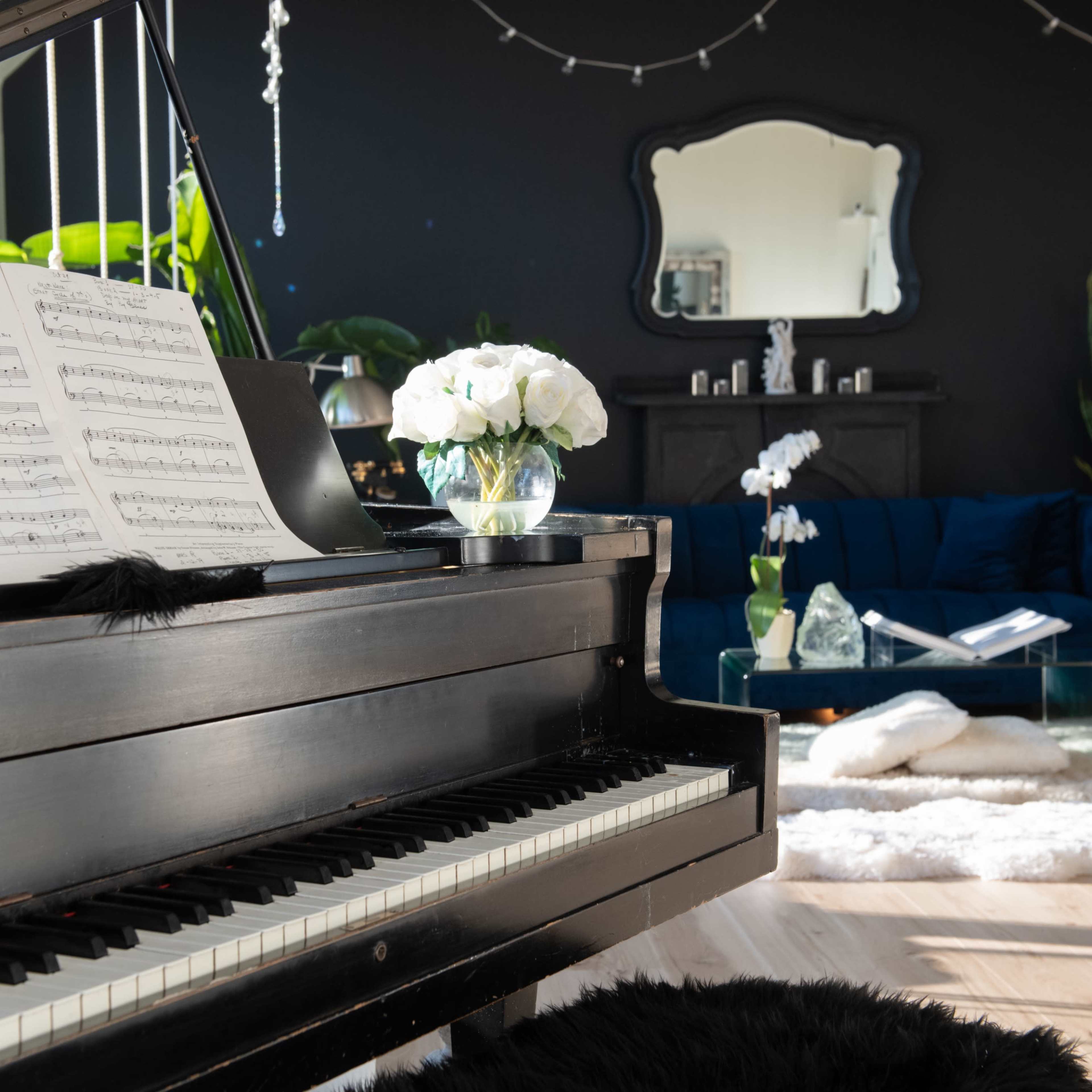A black piano with sheet music sits in a bright room featuring a large mirror, a blue couch, and various plants.