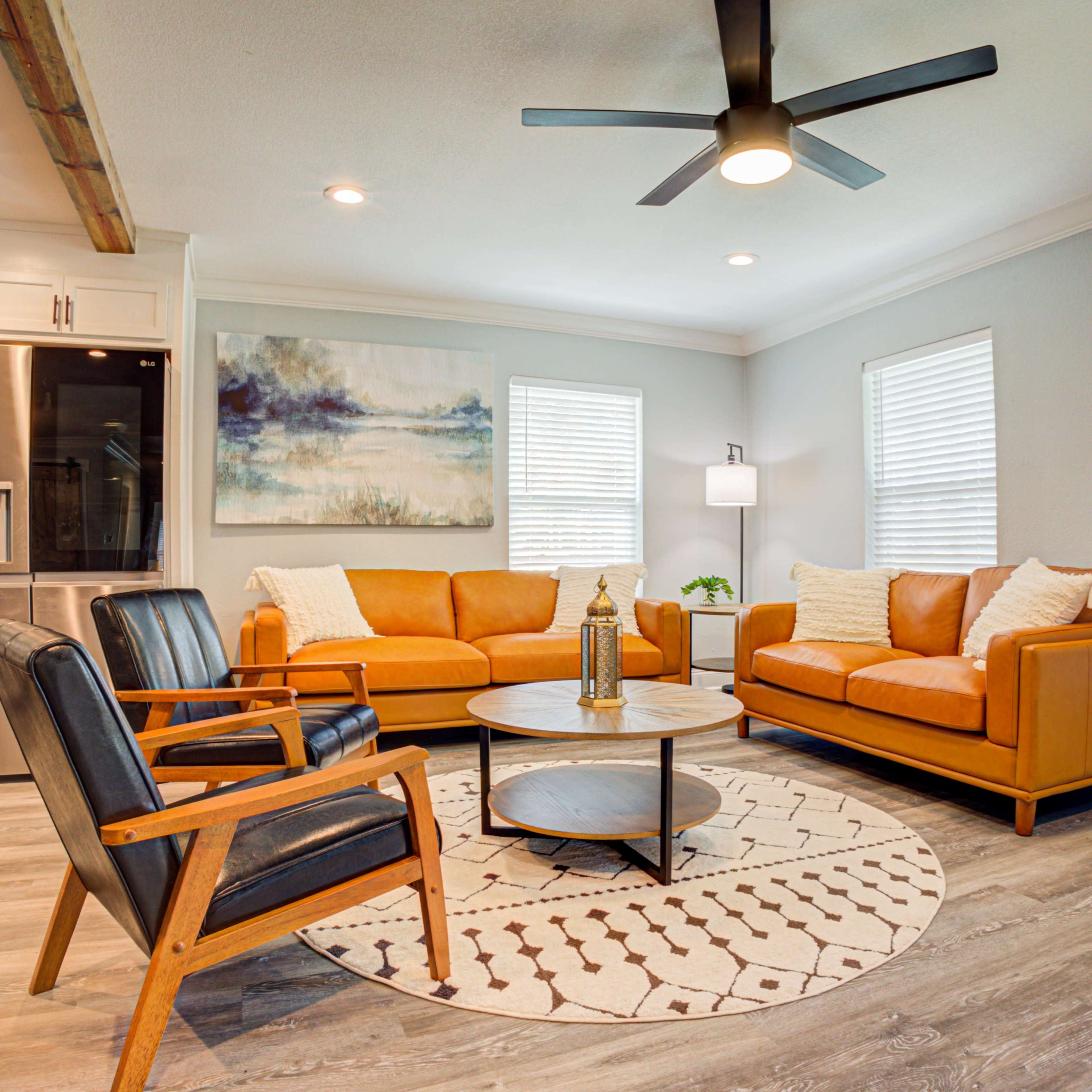A modern living room with two orange sofas, a round coffee table, and black armchairs, featuring a ceiling fan and light-colored walls.