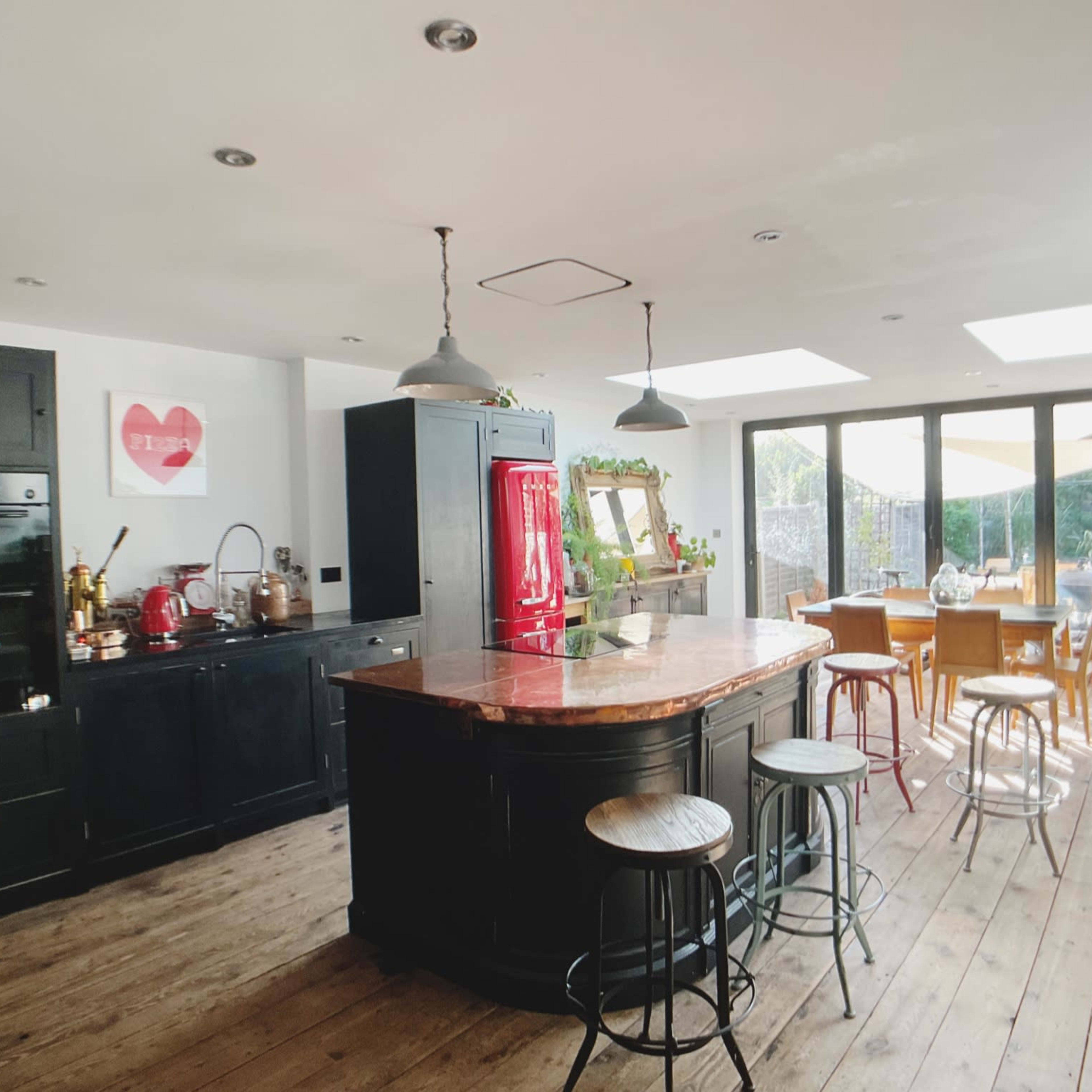 The kitchen features dark cabinetry, a large circular island with a copper countertop, and a dining area with wooden furniture illuminated by natural light from large windows.