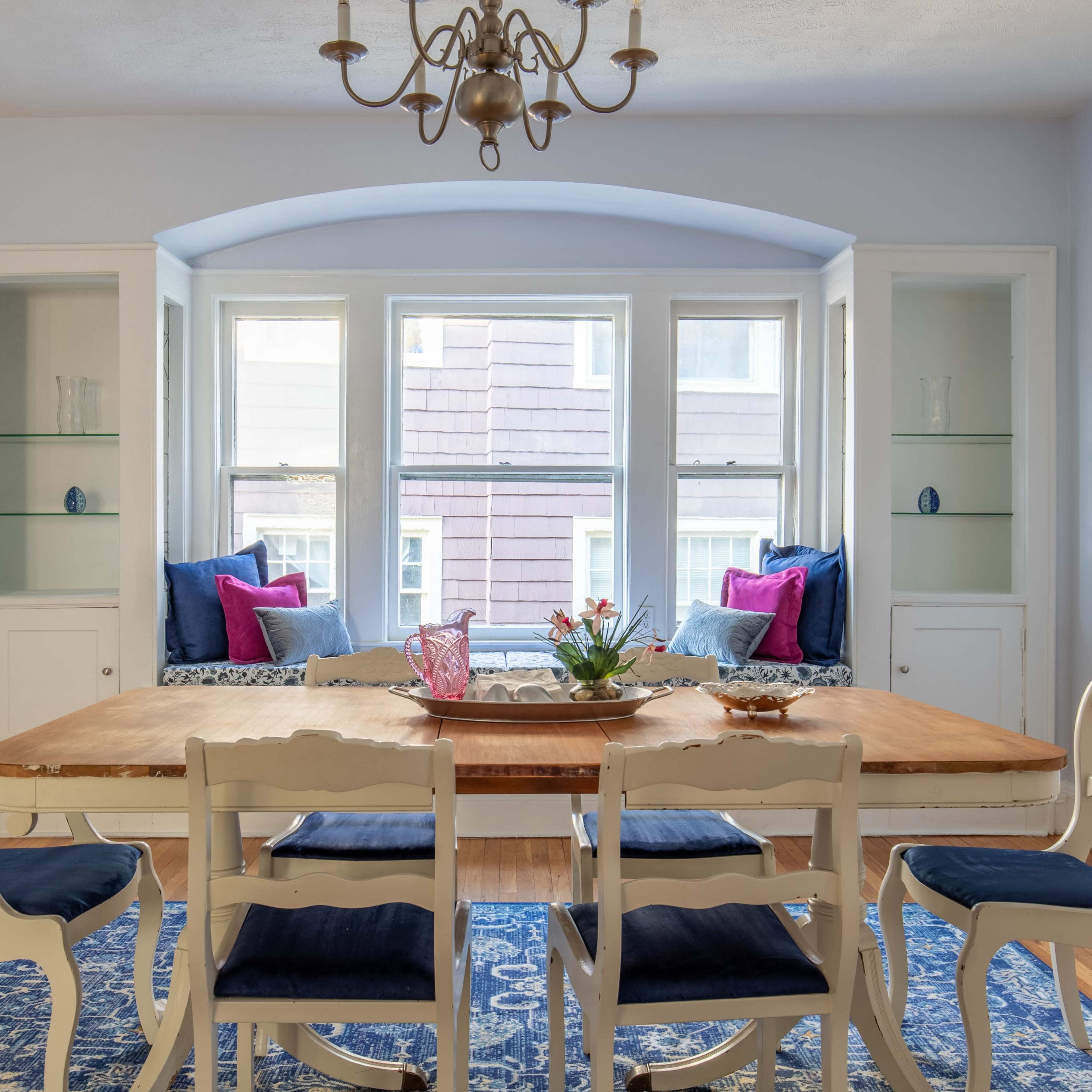 A dining room features a wooden table surrounded by six chairs, with a window nook on one side showcasing blue cushions and decorative elements.