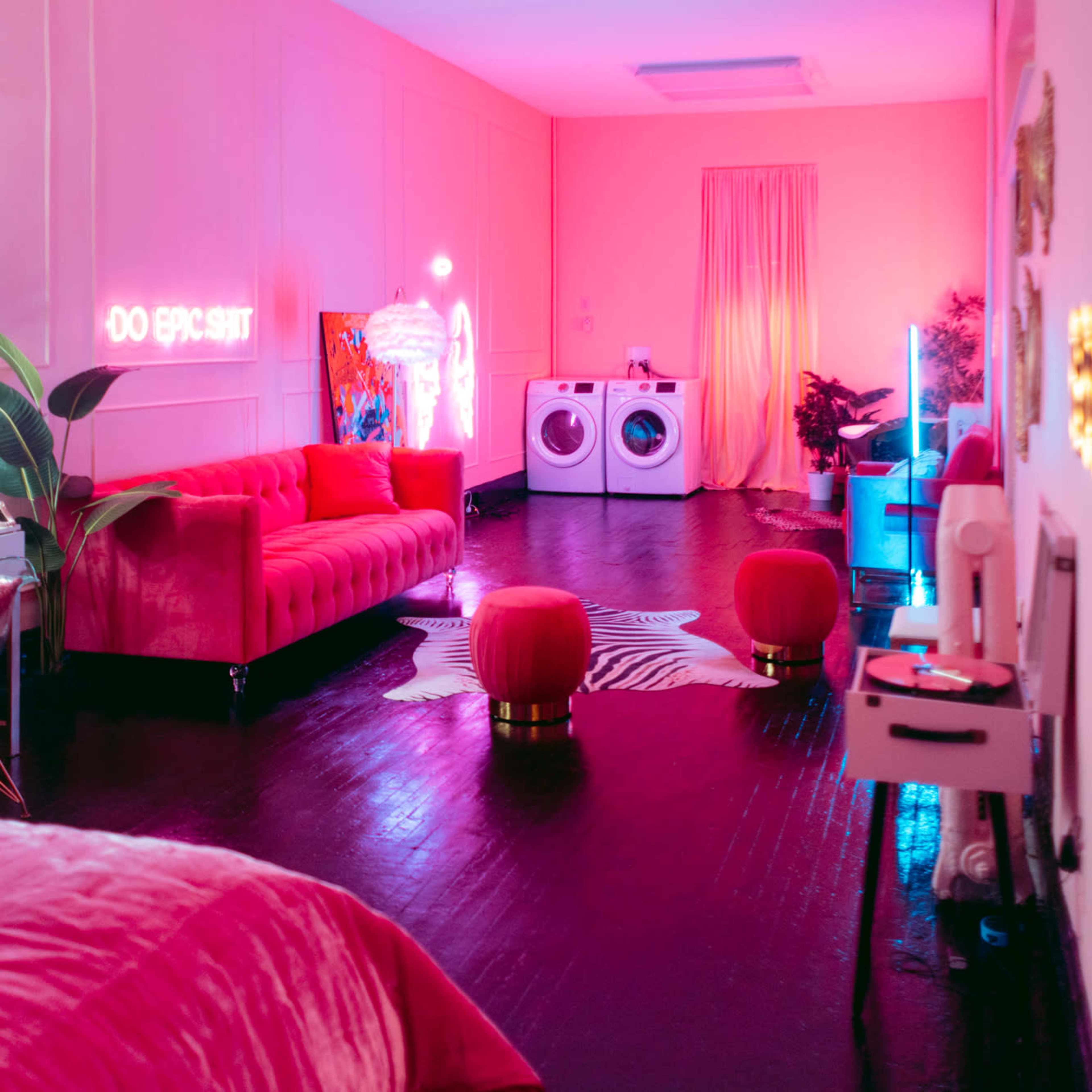 A brightly lit room features a pink couch, round poufs, a vanity area, and a washing machine, all set against a vibrant color scheme.