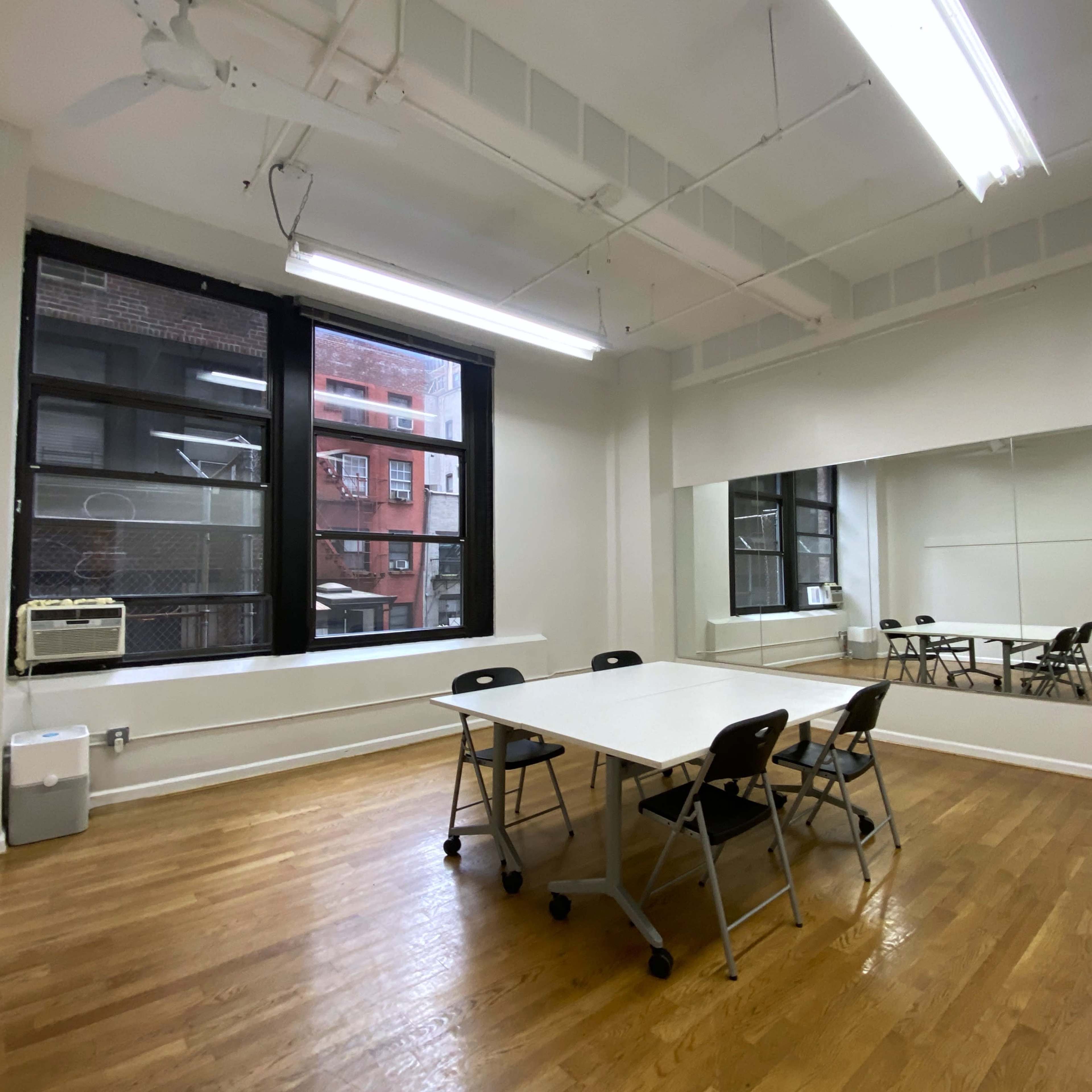 A sparsely furnished room with wooden floors, a large mirror on one wall, a table with chairs in the center, and windows that allow natural light.