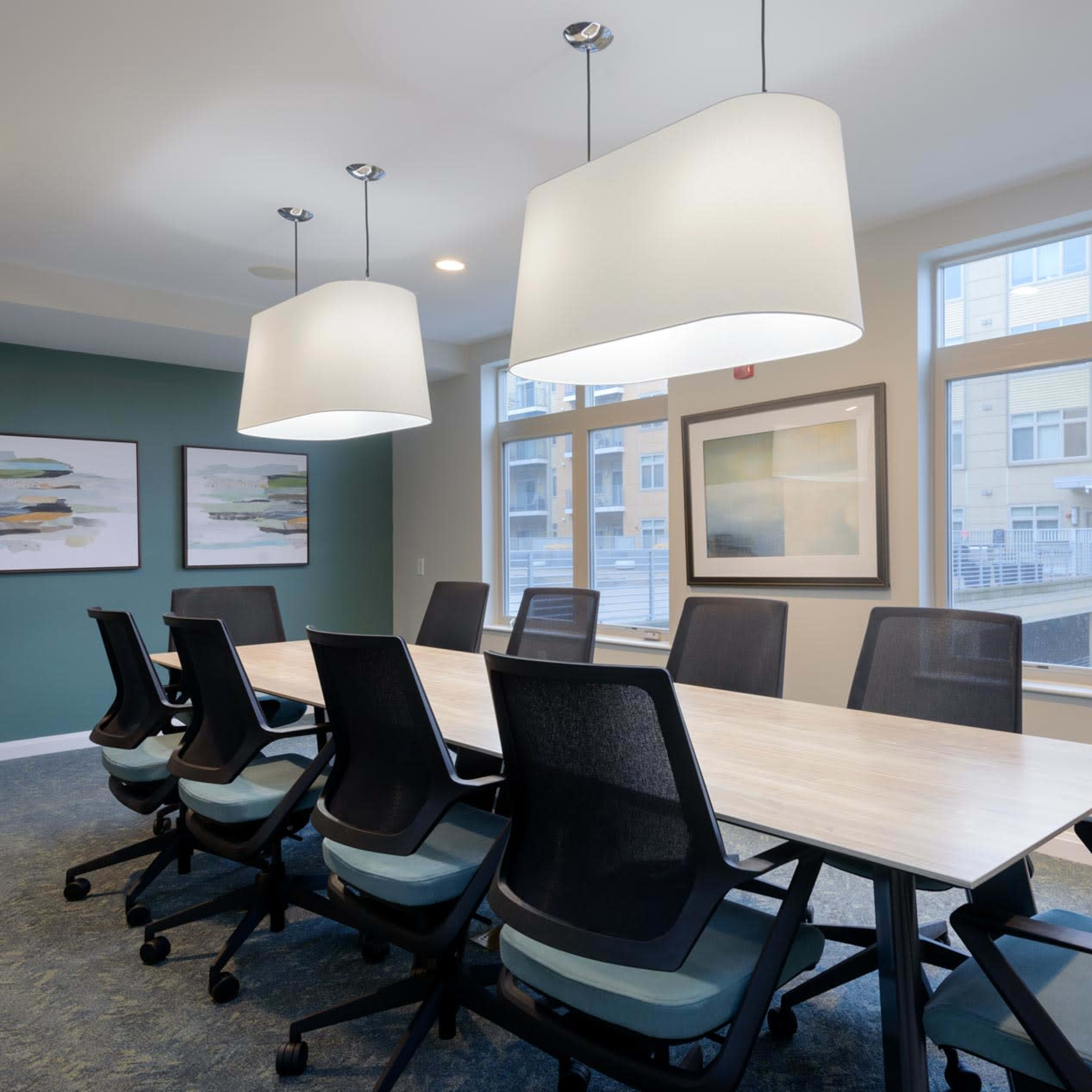 A modern conference room features a long table surrounded by eight ergonomic chairs, with large windows and two pendant lights overhead.