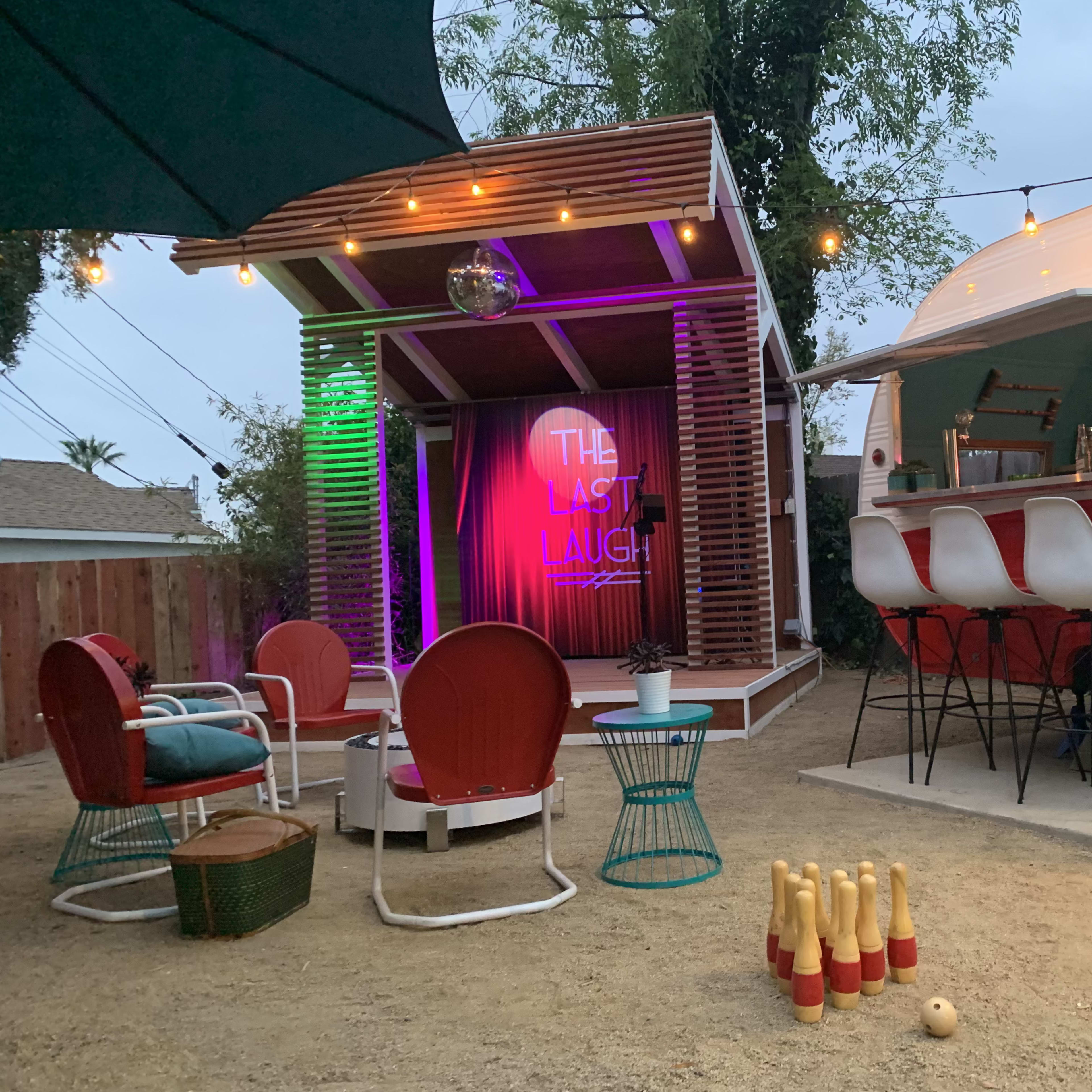 A cozy outdoor space featuring a retro trailer, a small stage with a colorful sign, and a bowling setup on the ground.