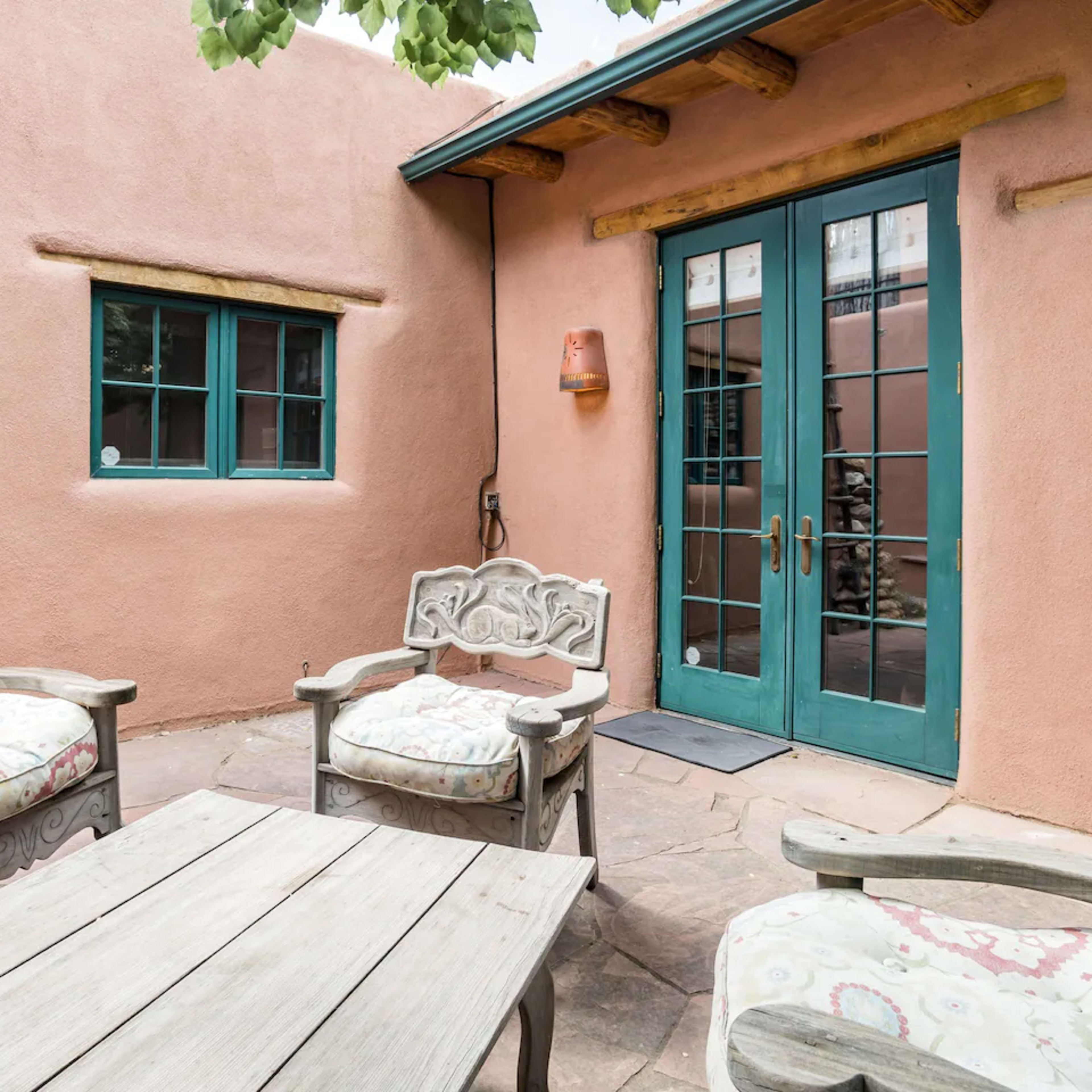 A courtyard features a wooden table surrounded by four chairs, with a ladder against the pink adobe wall and large windows leading indoors.