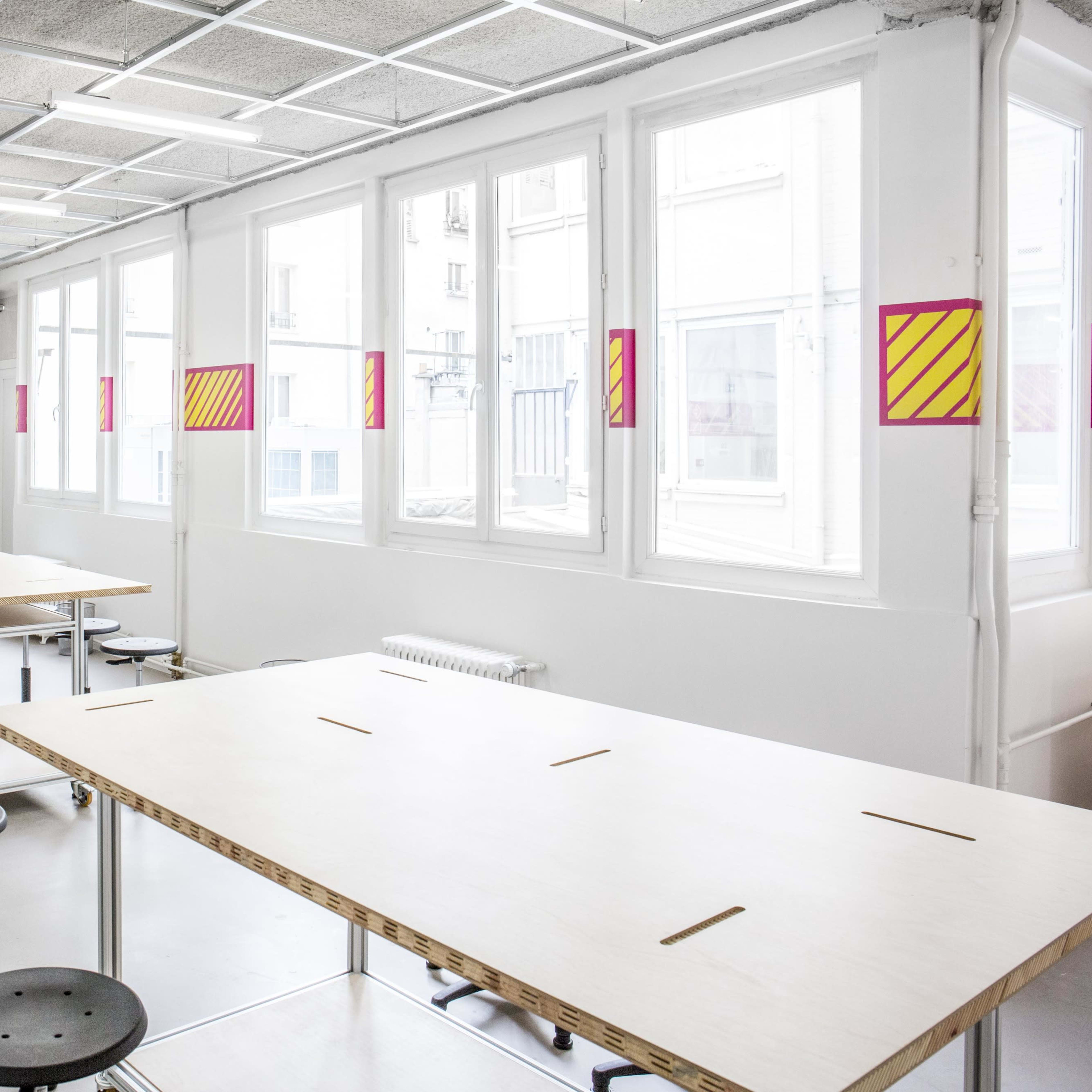 A bright, empty classroom with several wooden tables and rolling stools arranged neatly under large windows.