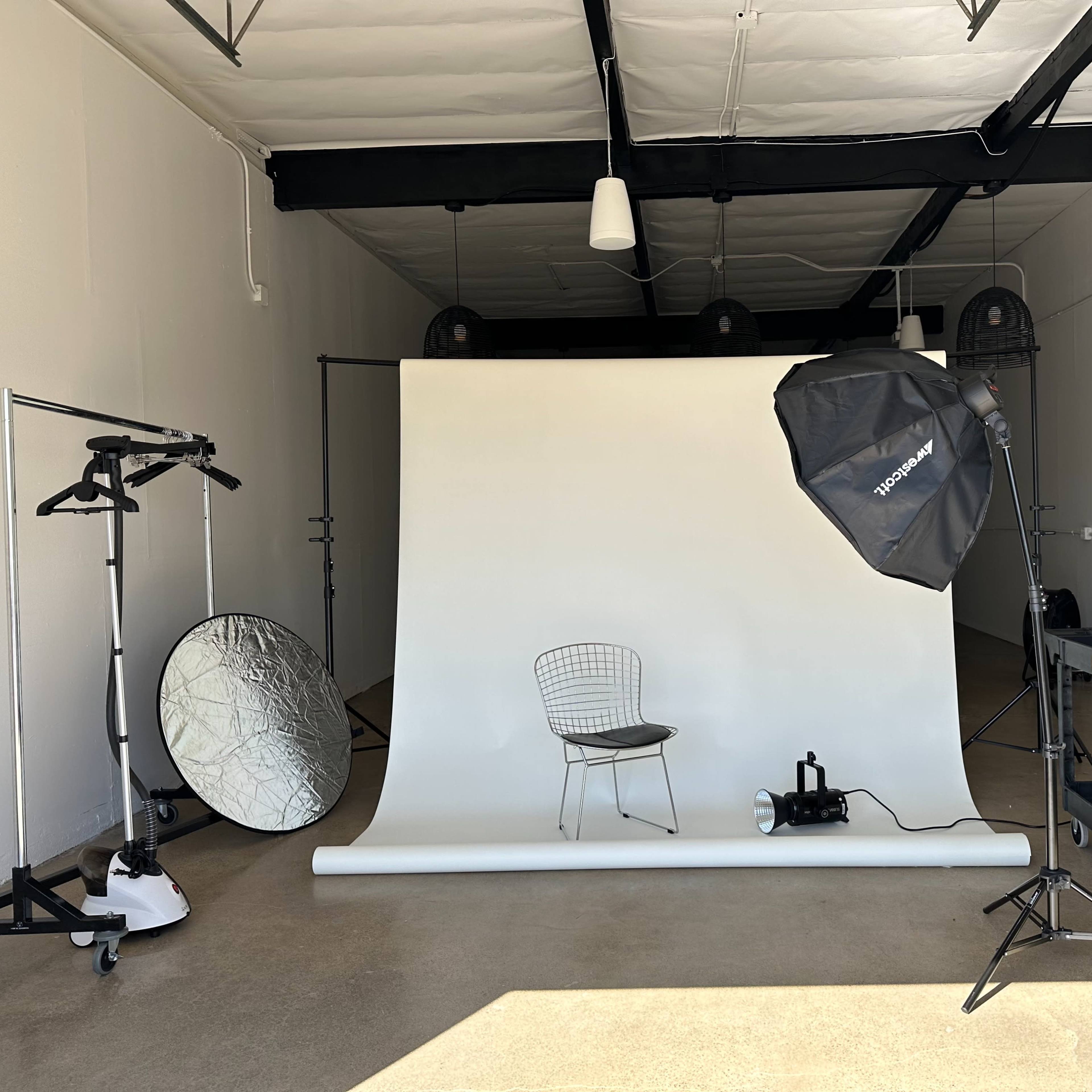 A photography studio featuring a white backdrop, a single chair, and lighting equipment.