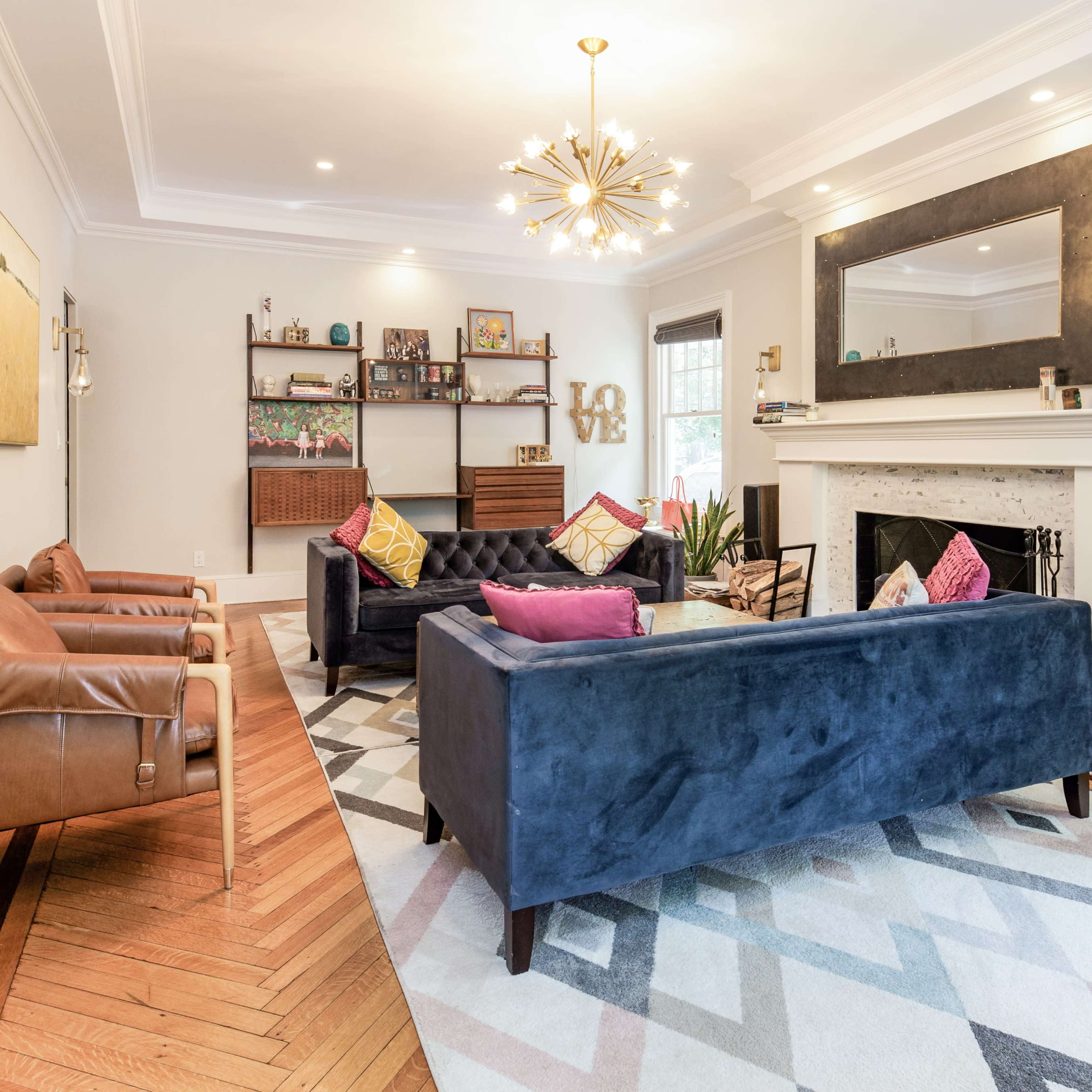 A living room features two couches, armchairs, a coffee table, and a decorative fireplace, all arranged on a geometric patterned rug.