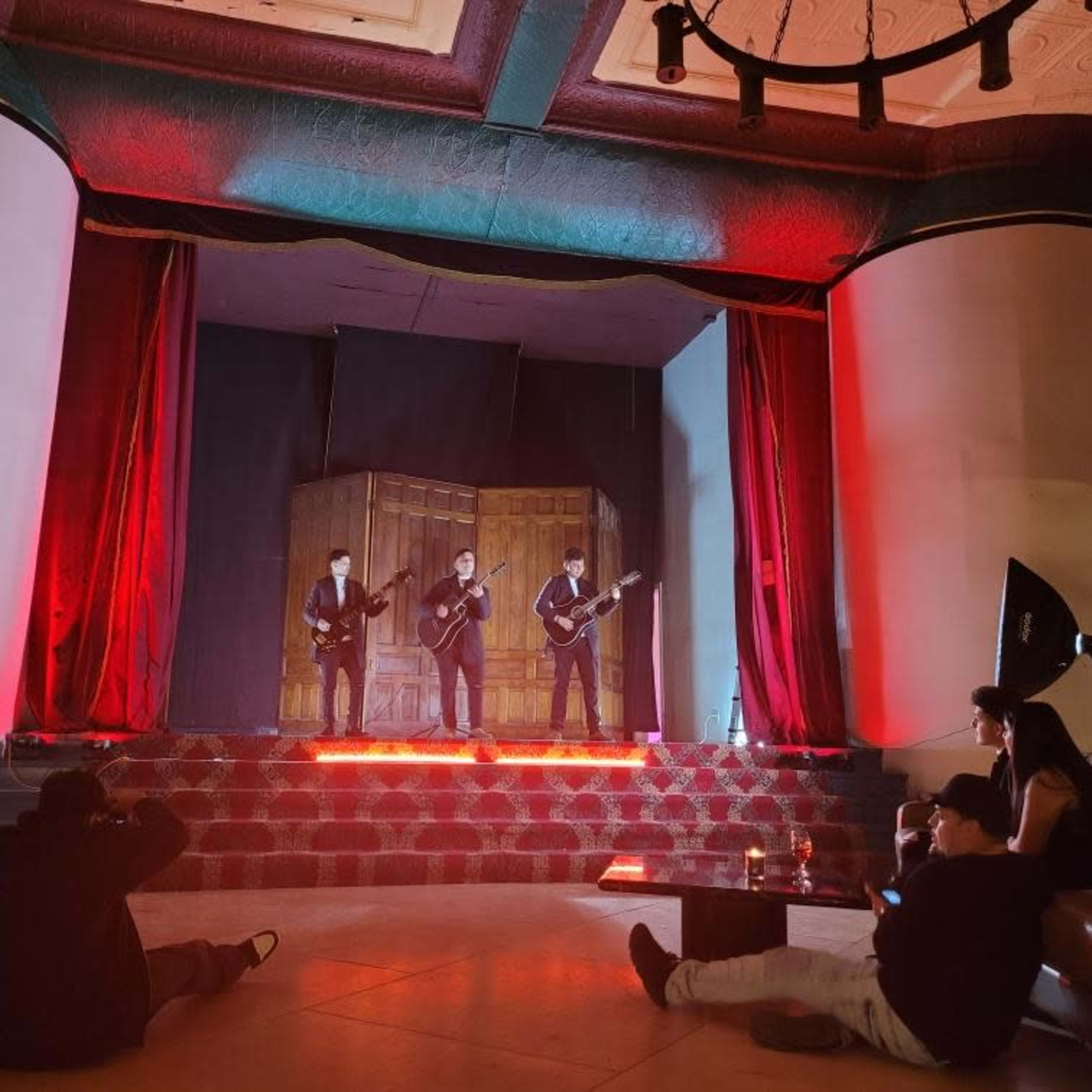 Three musicians perform on stage in front of a small audience in a venue with red lighting and a dark backdrop.