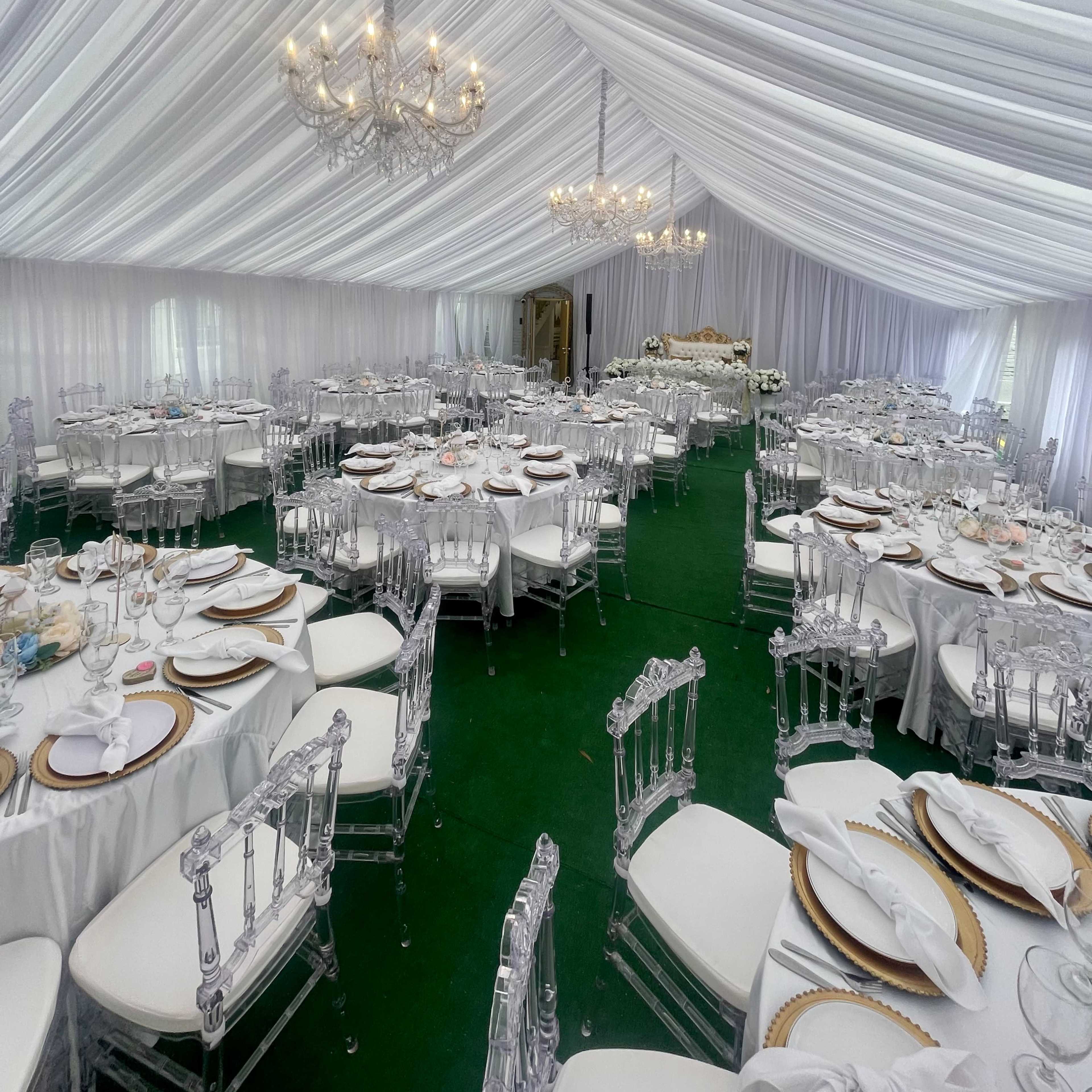 A large event tent is set up with white draped ceilings, round tables dressed in white linens, and clear acrylic chairs arranged neatly on green carpet.