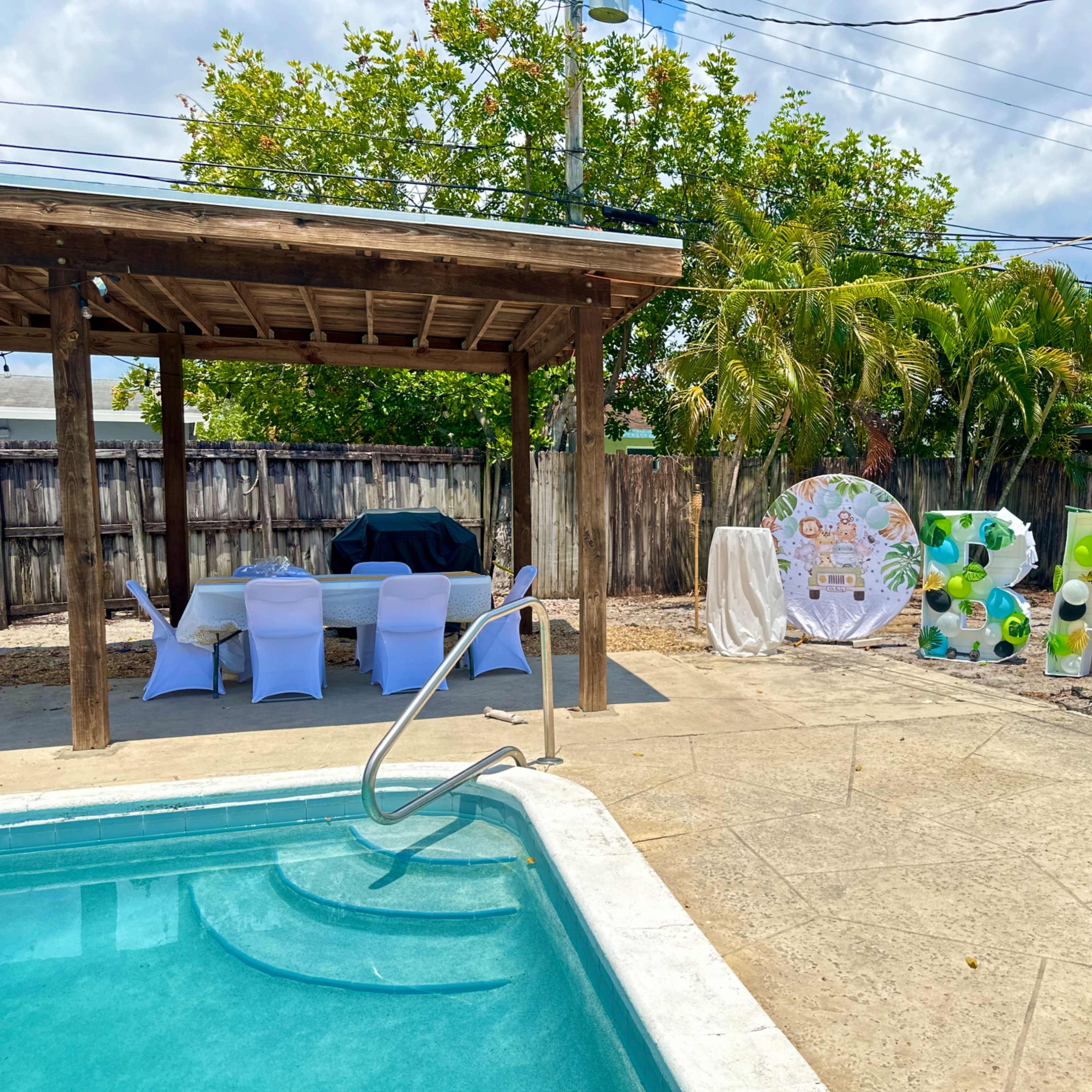 A backyard pool area with a wooden gazebo, seating arrangements under it, and a large decorative sign spelling "BABY" adorned with colorful balloons.