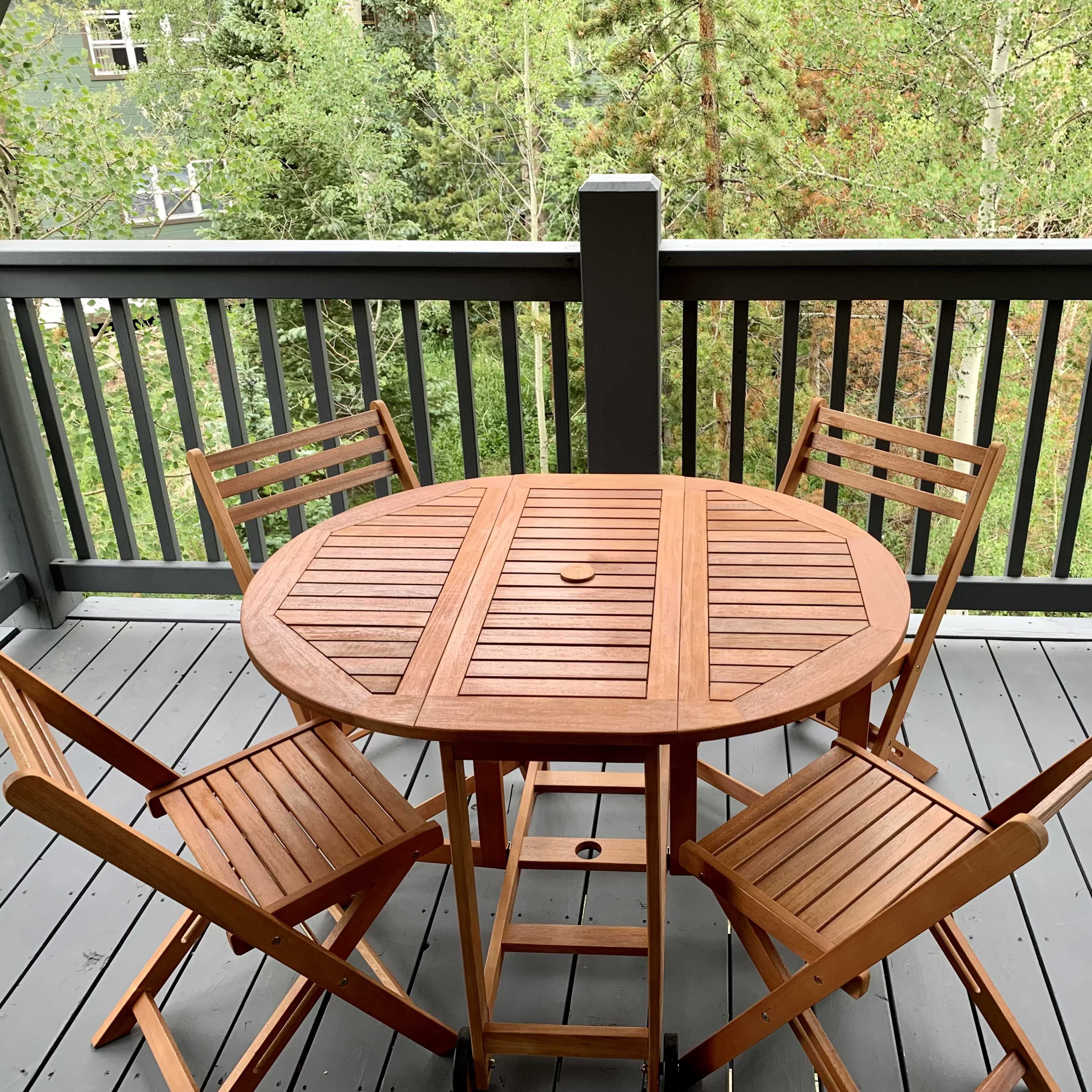 A round wooden table surrounded by four folding chairs is positioned on a covered balcony overlooking green trees.