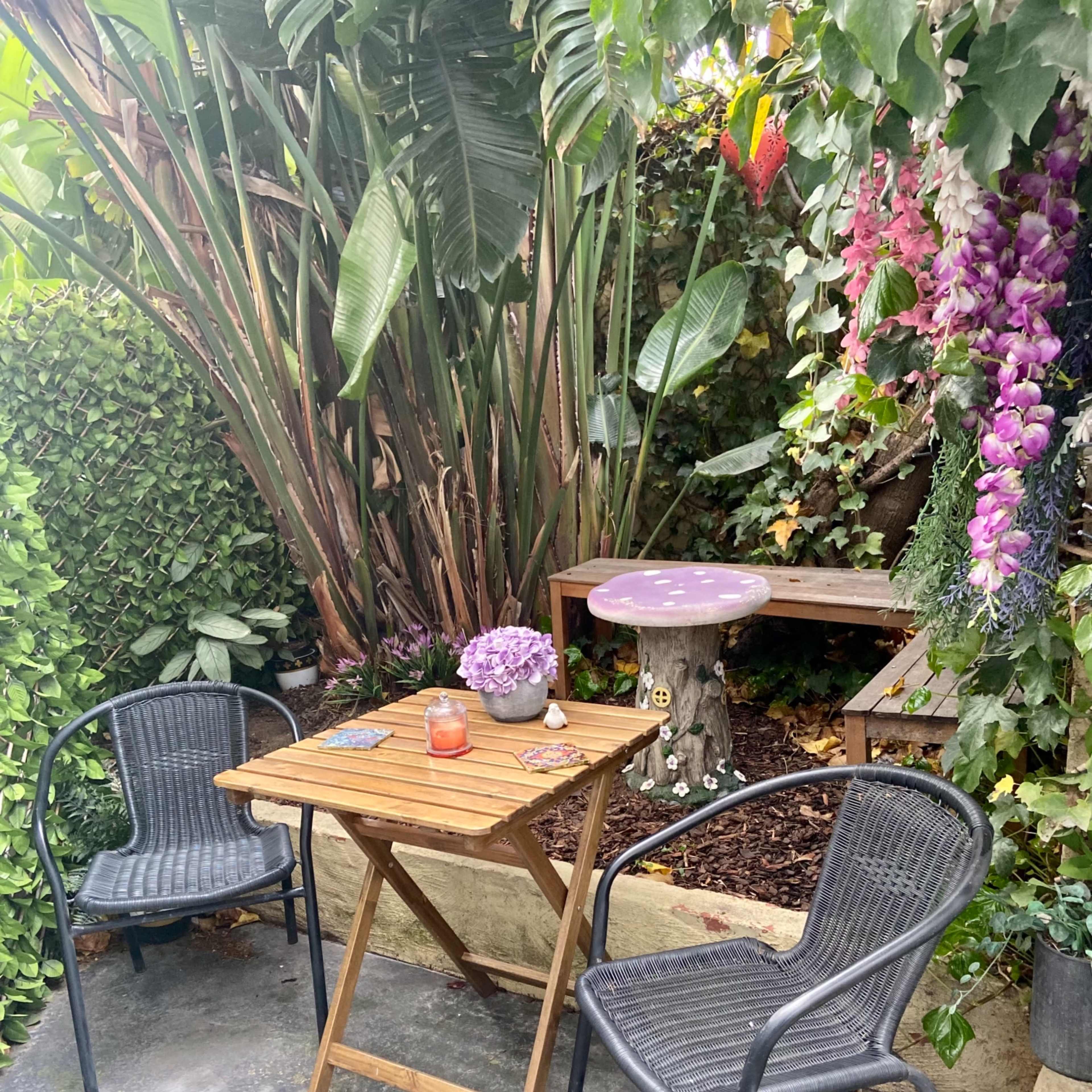 A small wooden table with two chairs is set up in a lush garden filled with tropical plants and flowers.