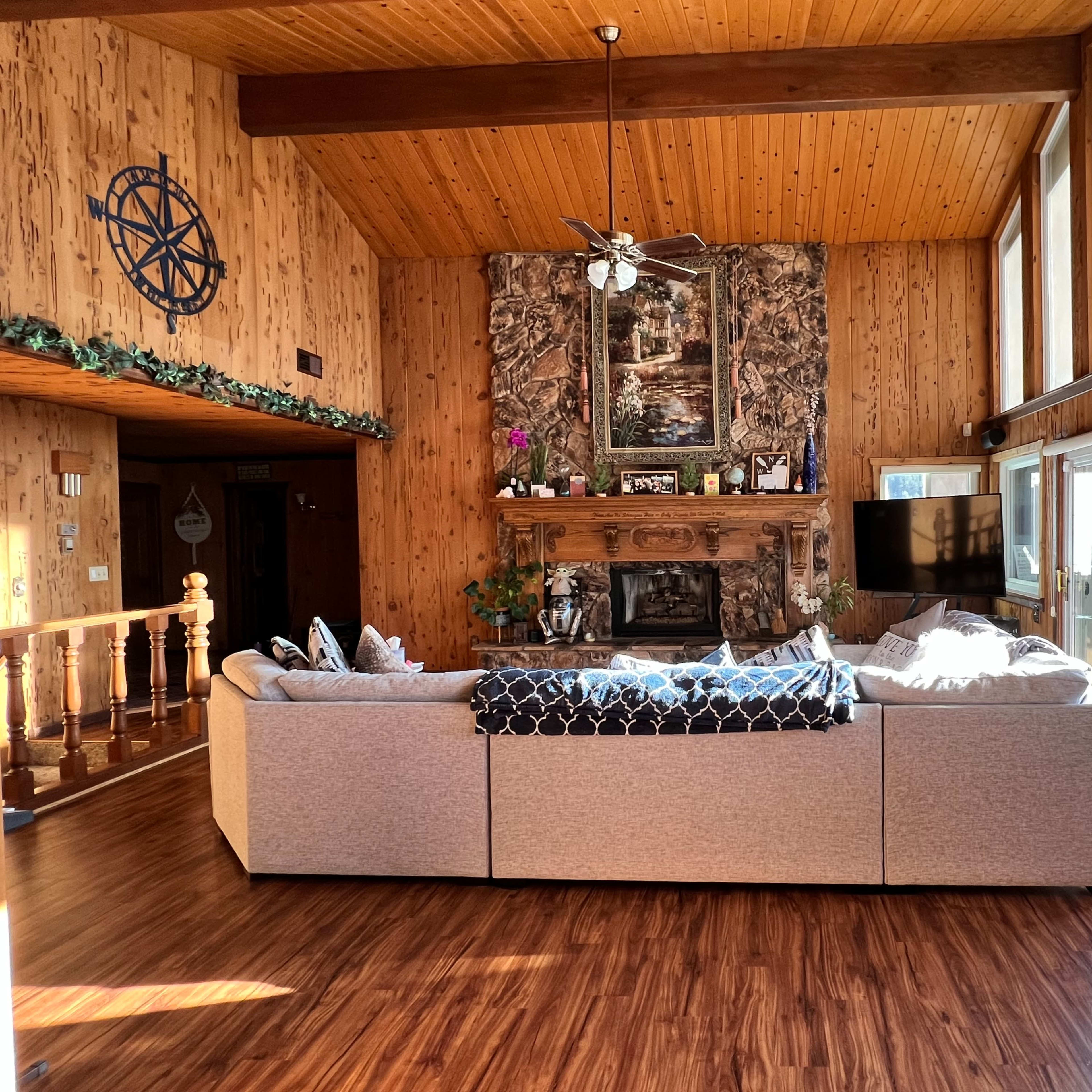 A spacious living room with wooden walls, a large stone fireplace, and a sectional sofa facing a wall of windows.