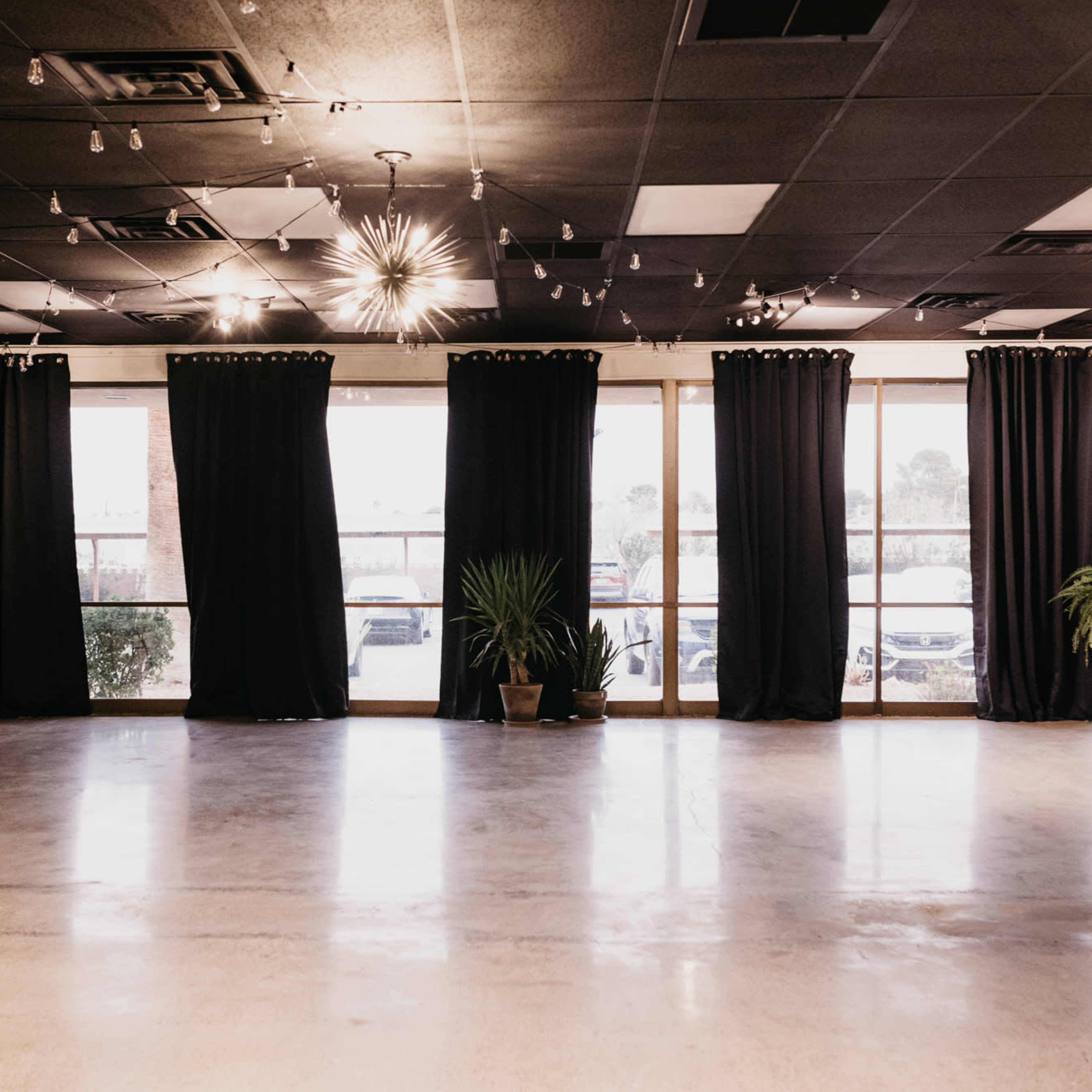 An empty event space with black curtains covering large windows, a teal couch against a wall, and potted plants in the corners.