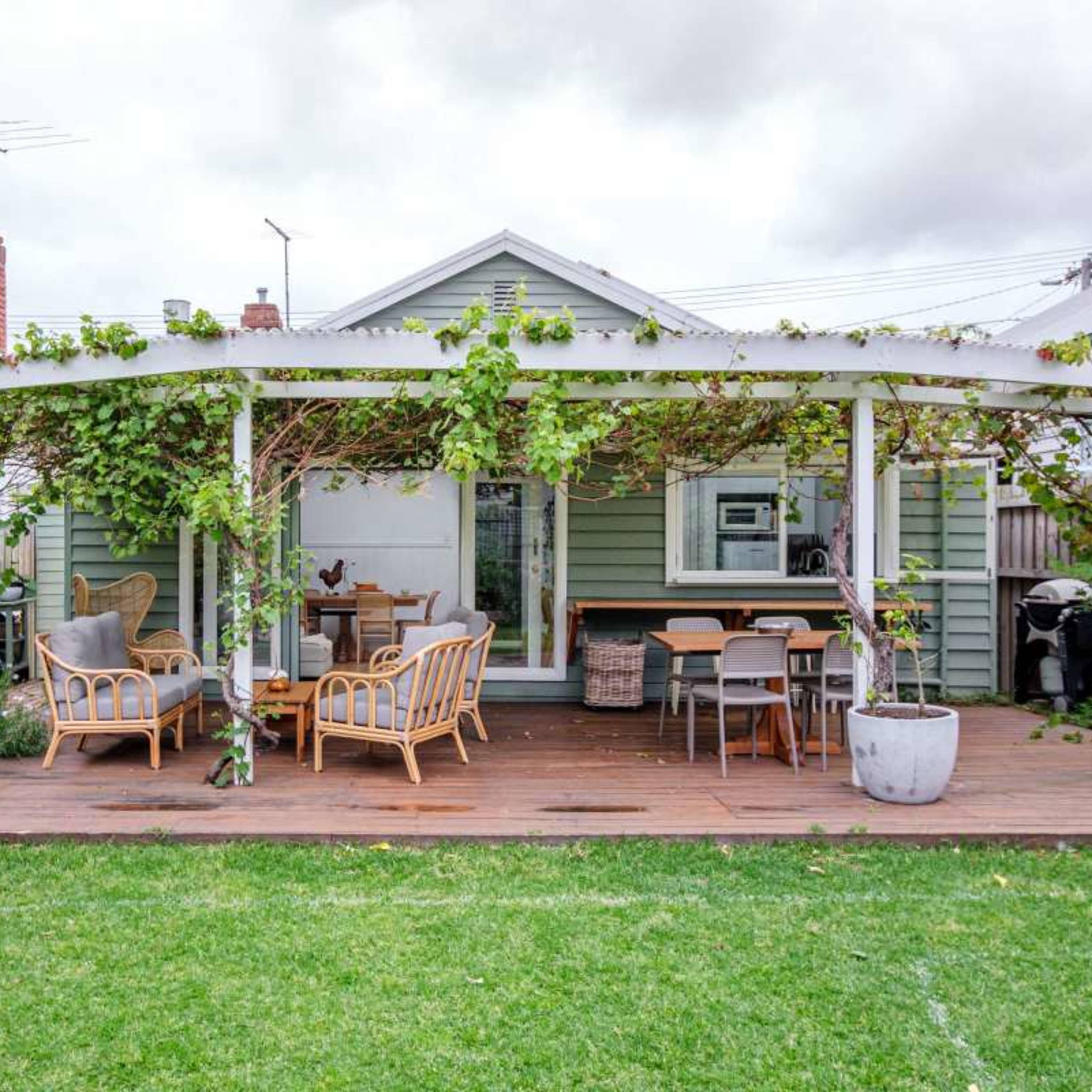 A wooden deck with seating and a dining area is covered by a pergola adorned with greenery, leading to a modest house.