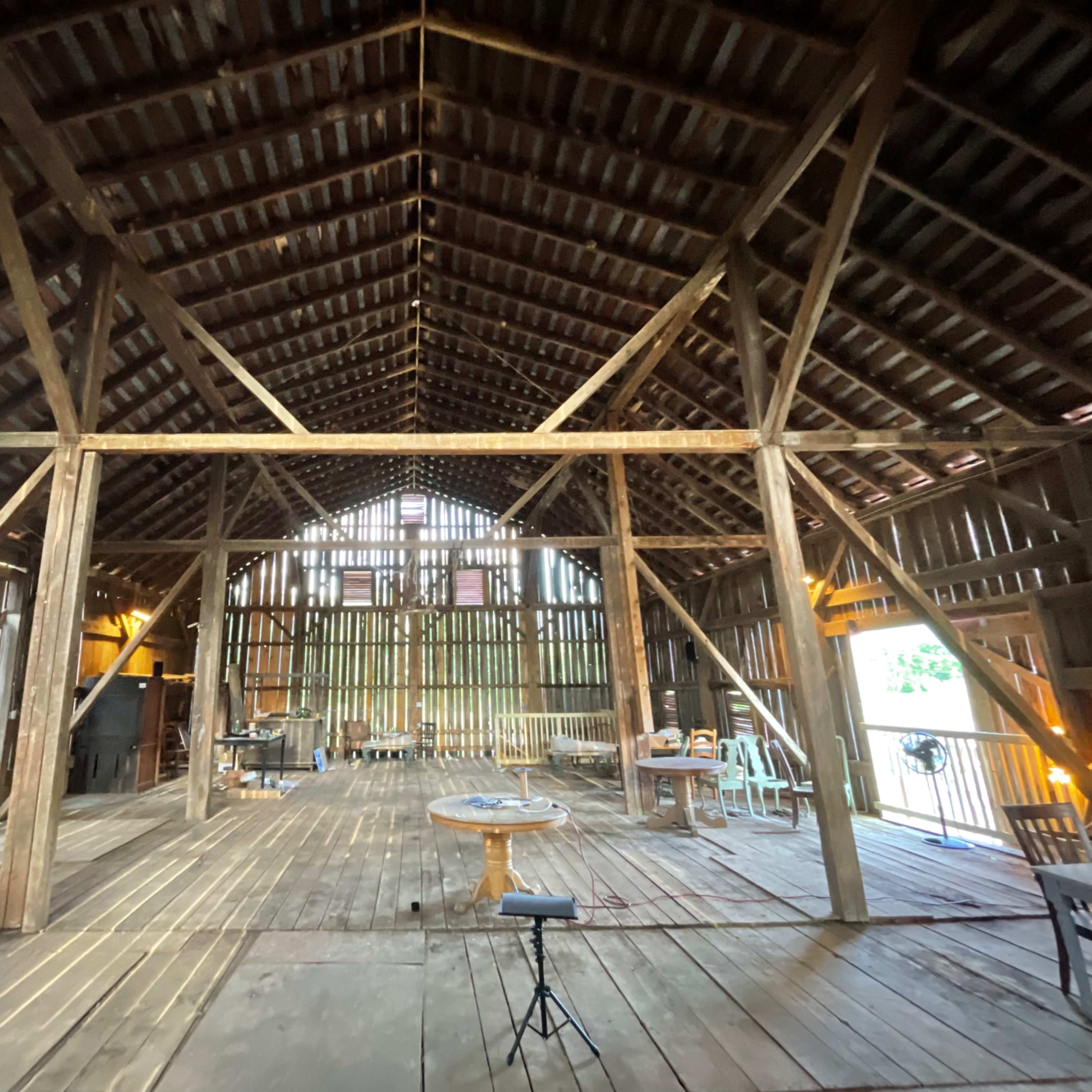 The interior of a spacious, rustic barn with exposed wooden beams, various furniture pieces, and tools scattered across the wooden floor.