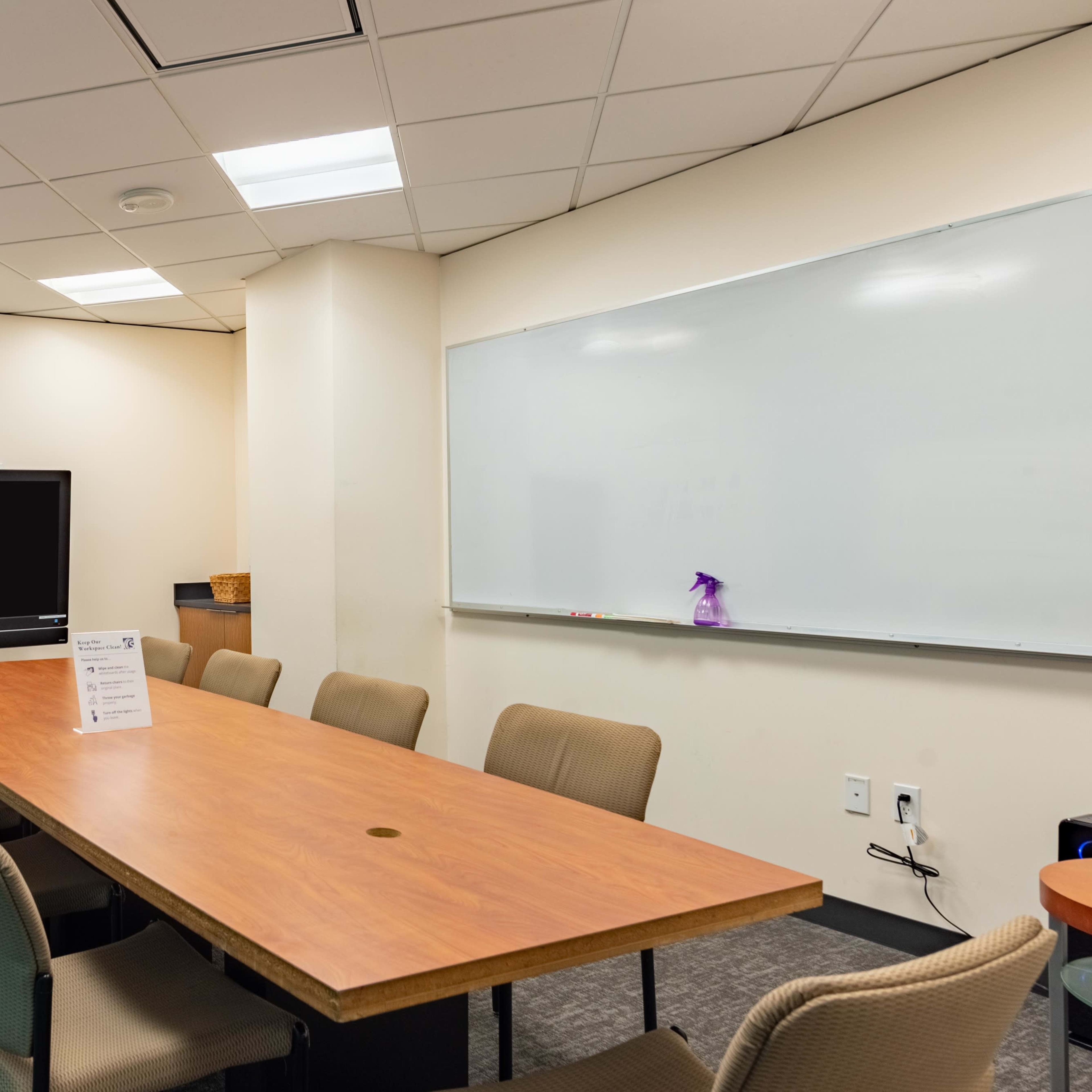 A conference room features a long wooden table surrounded by beige chairs, a large screen on one wall, and a whiteboard on another.