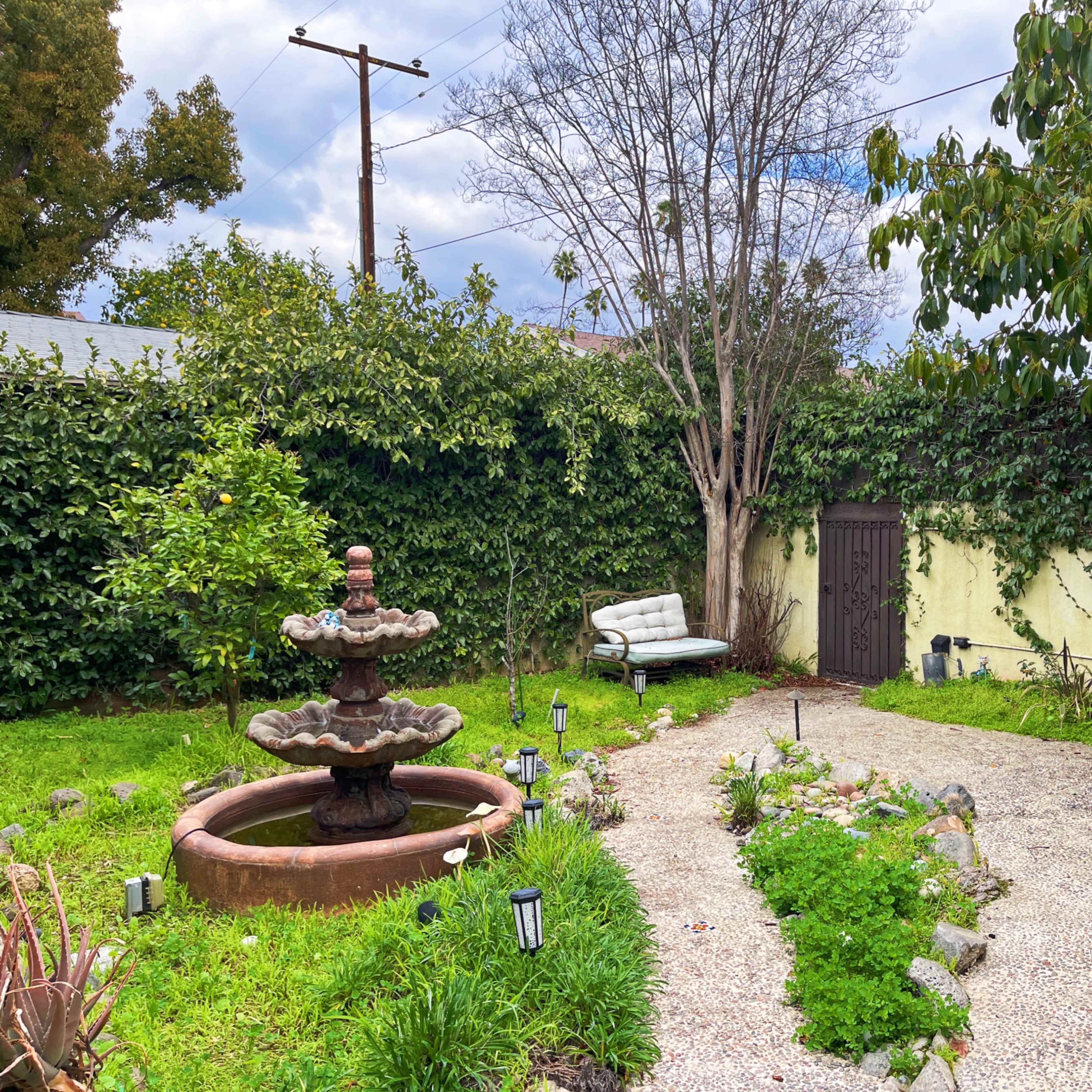 A small garden features a fountain, a bench, and surrounding greenery with a gravel path.