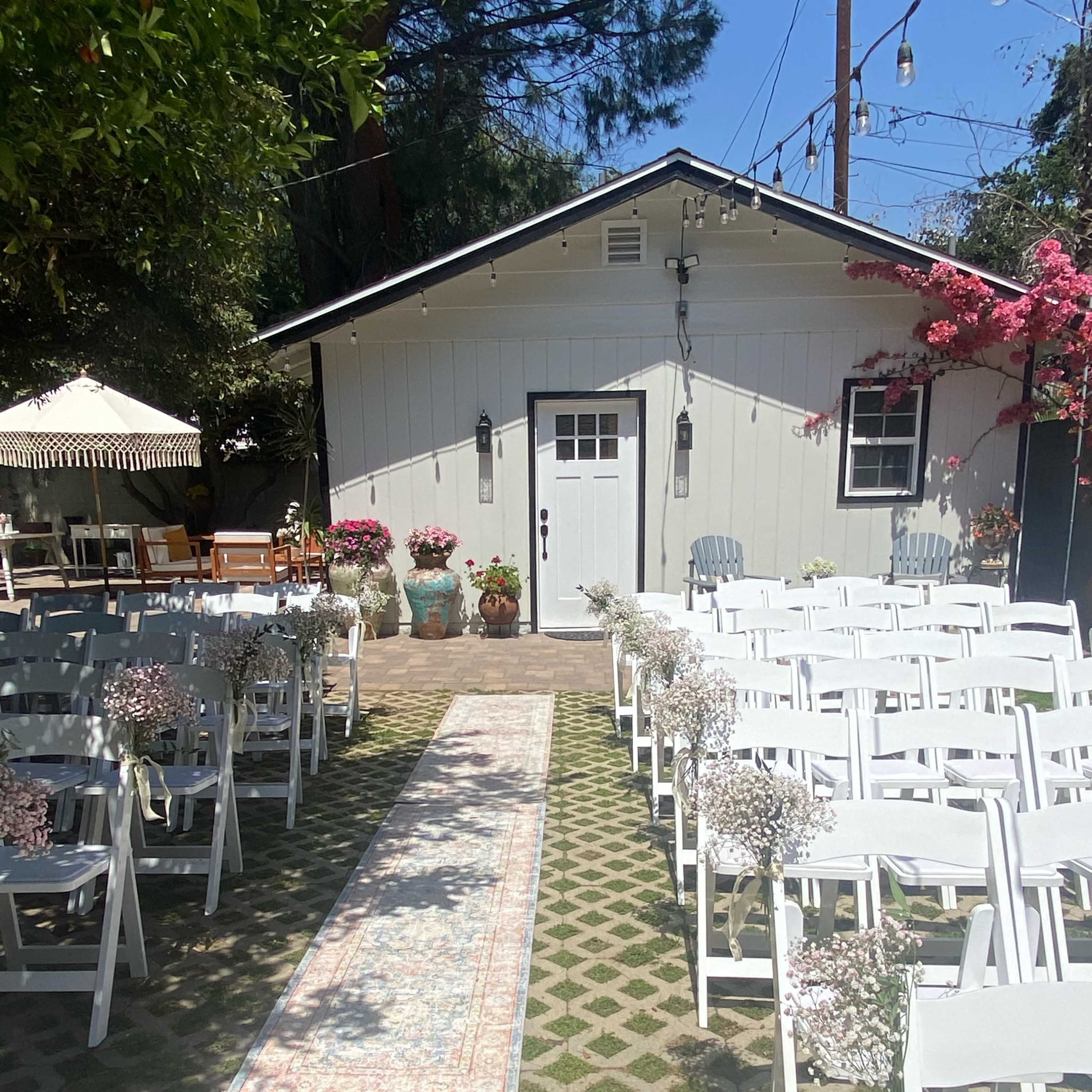 The scene shows a wedding setup with white chairs arranged in rows on a stone pathway leading to a white building, adorned with potted flowers and a decorative rug.