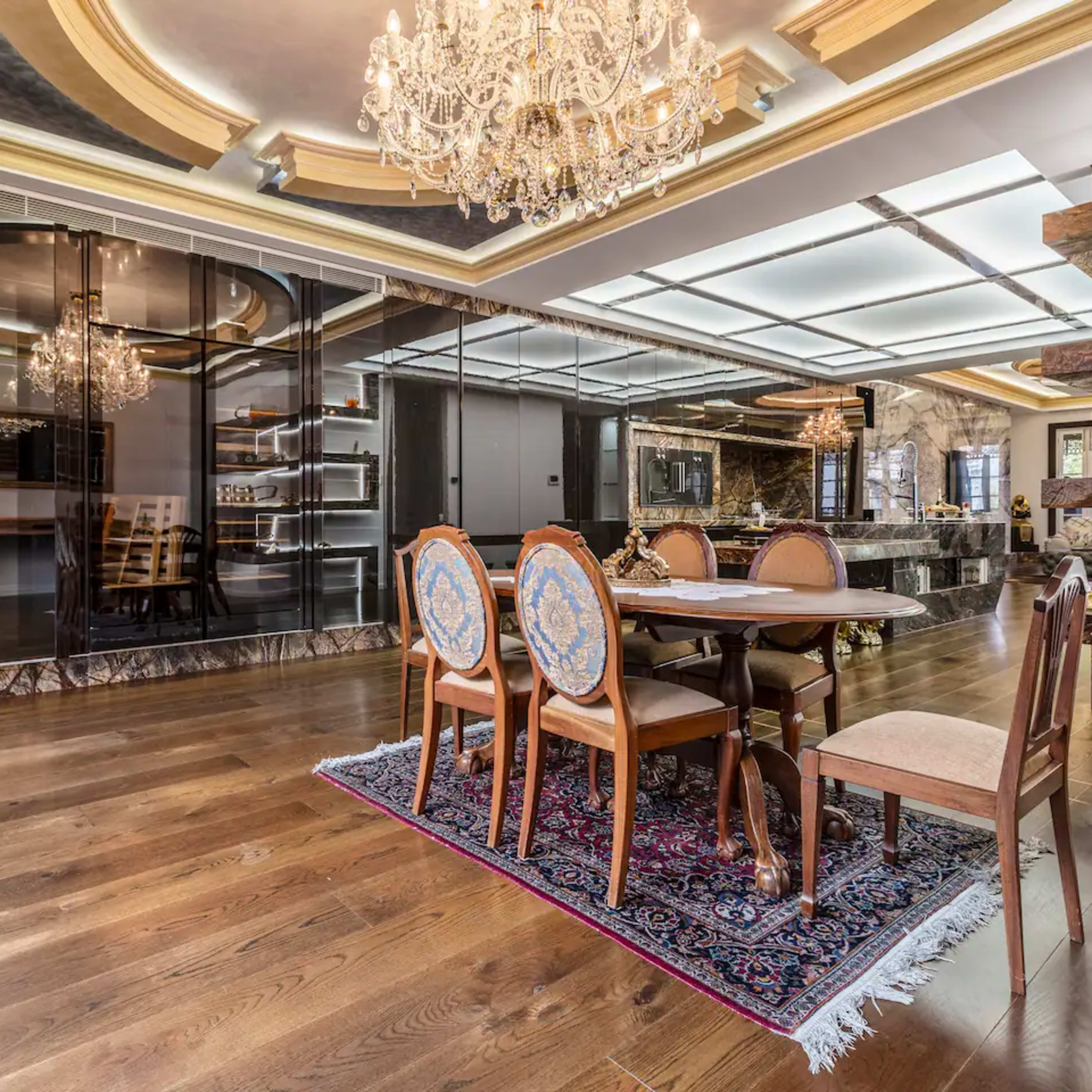 A luxurious dining area with a large table surrounded by elegant chairs, a chandelier overhead, and a marble-finished kitchen in the background.