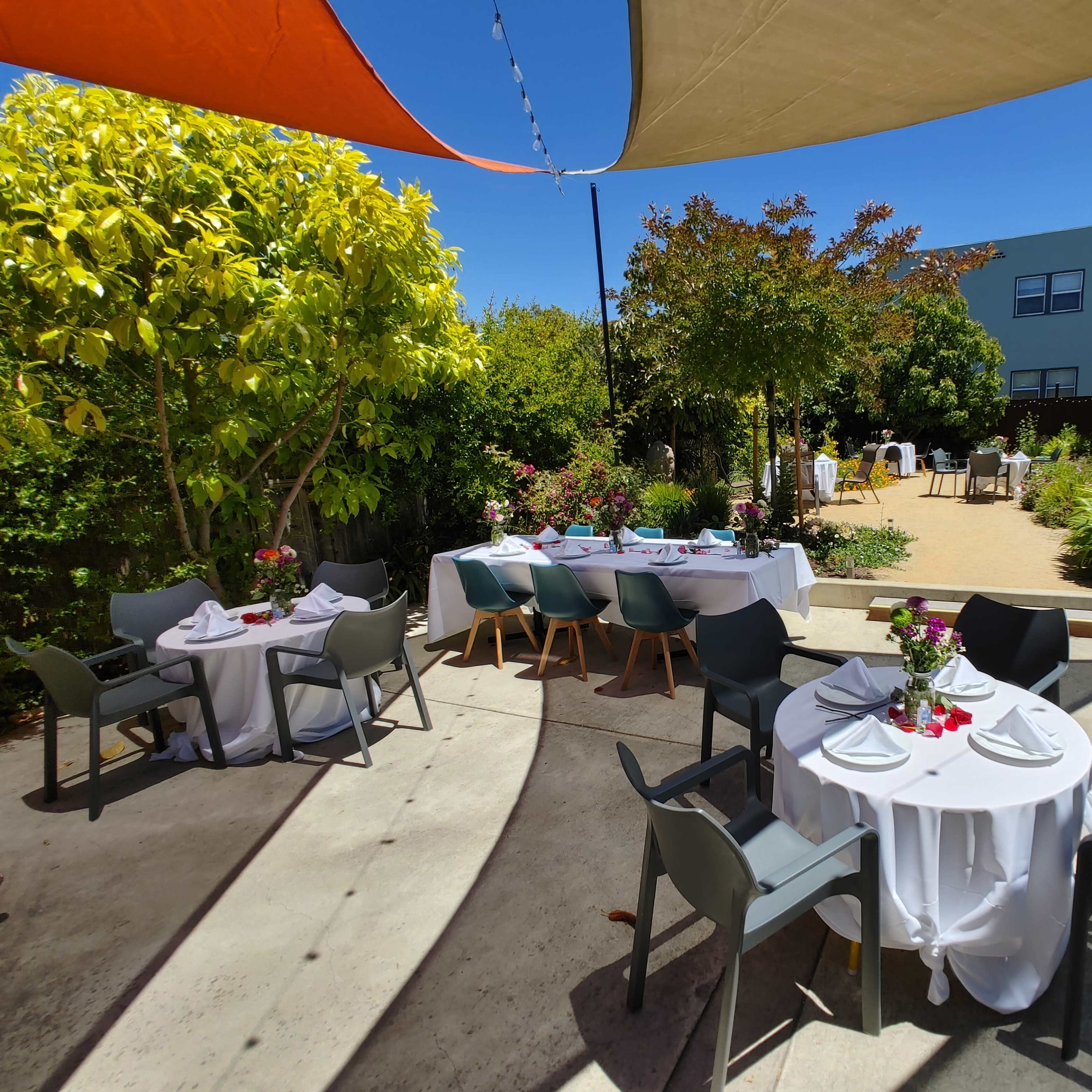 A sunny outdoor dining area features tables set with white tablecloths and flowers, surrounded by greenery and a building in the background.