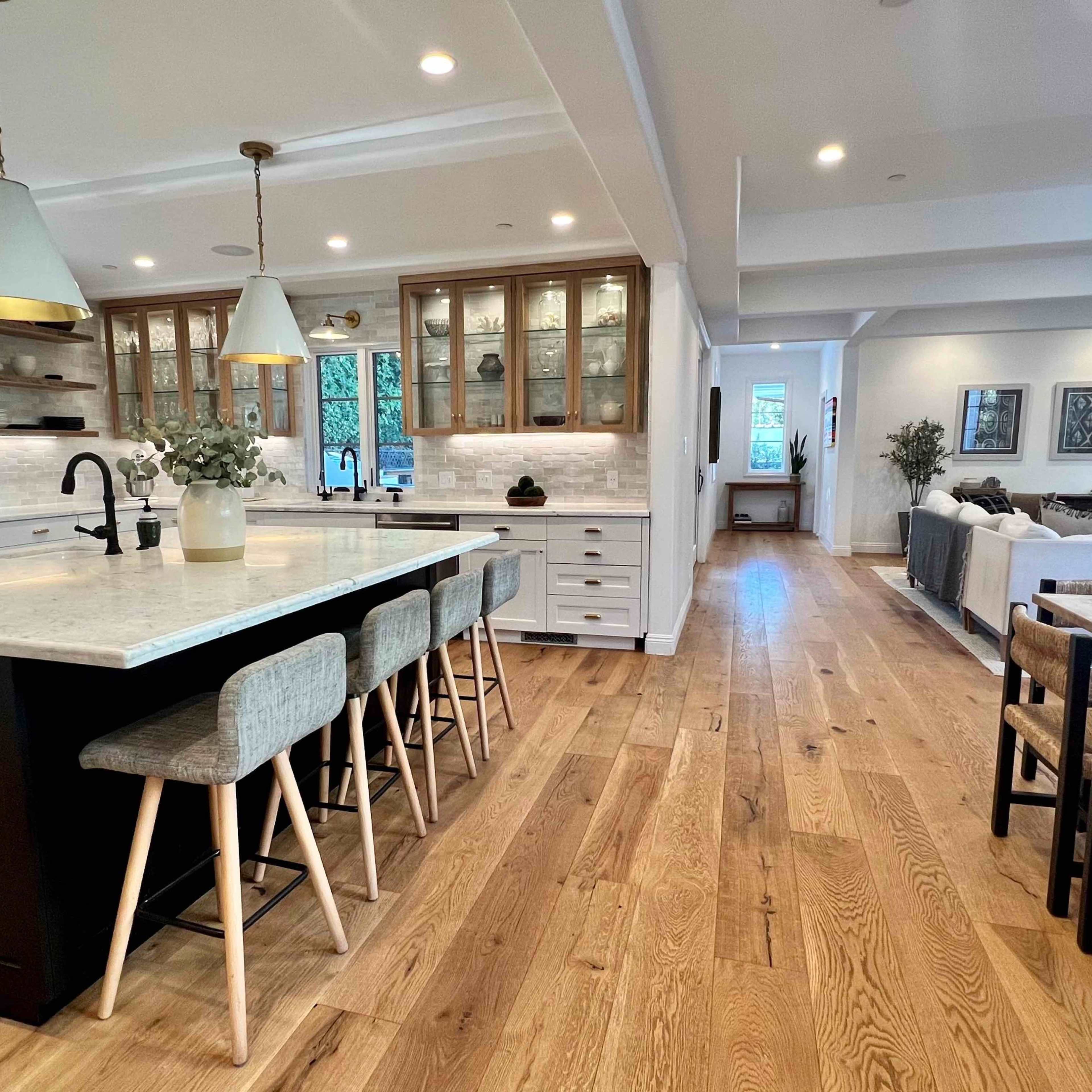 A modern kitchen with a large marble island, wooden flooring, and an open living area in the background.