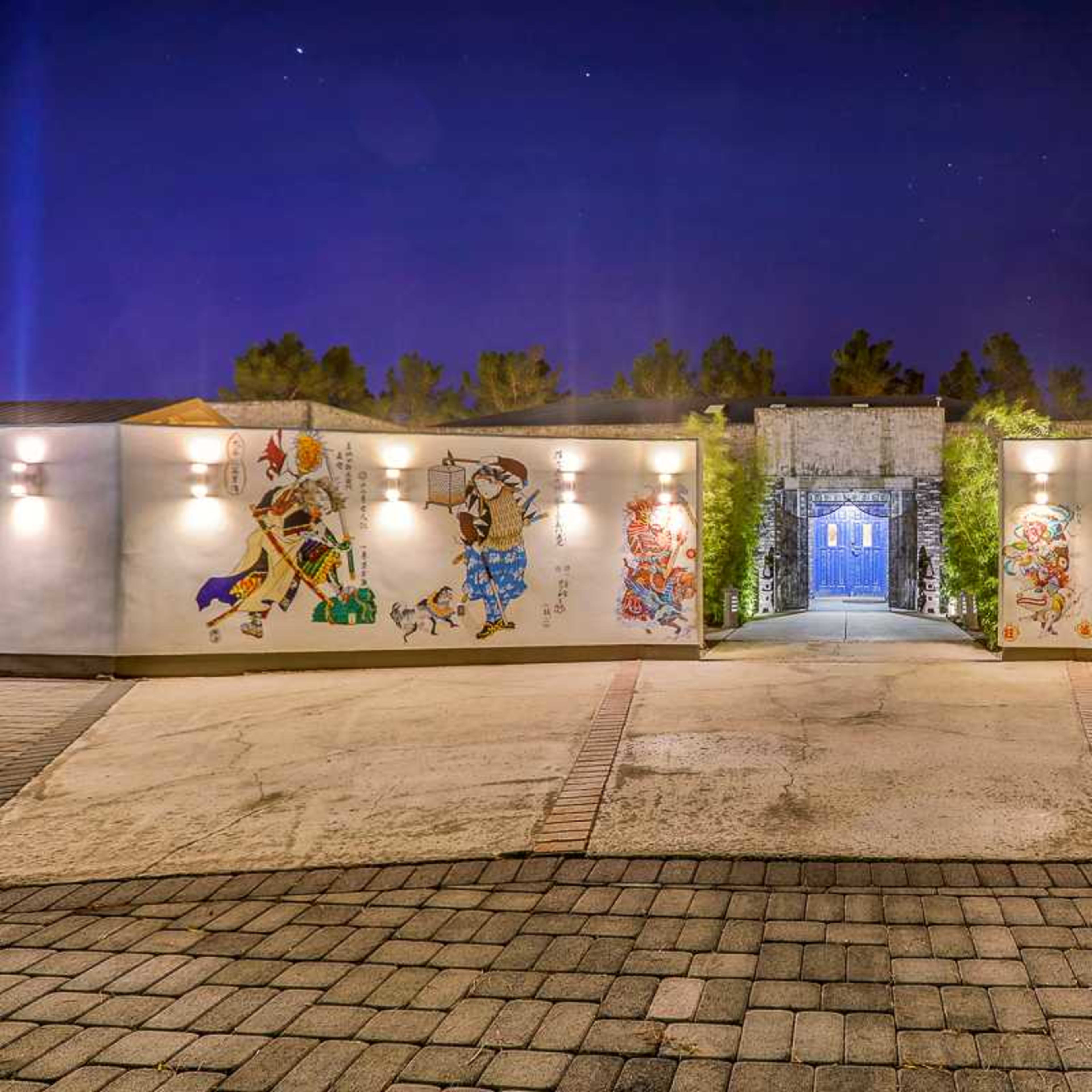 A pathway leading to a blue door framed by illuminated walls featuring colorful murals and surrounded by trees at night.