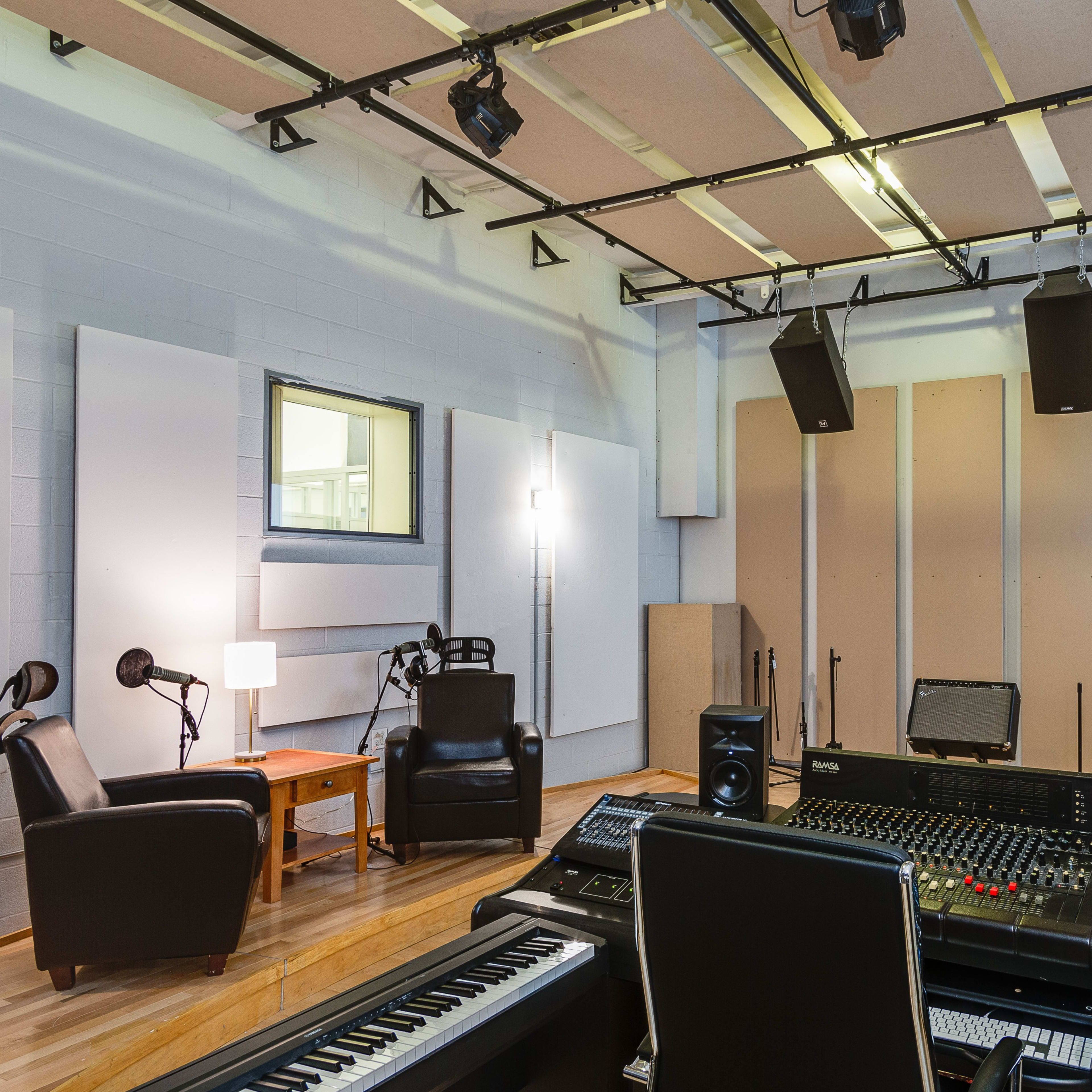 A sound recording studio featuring a control console, acoustic panels on the walls, two chairs, a side table, and overhead speakers.