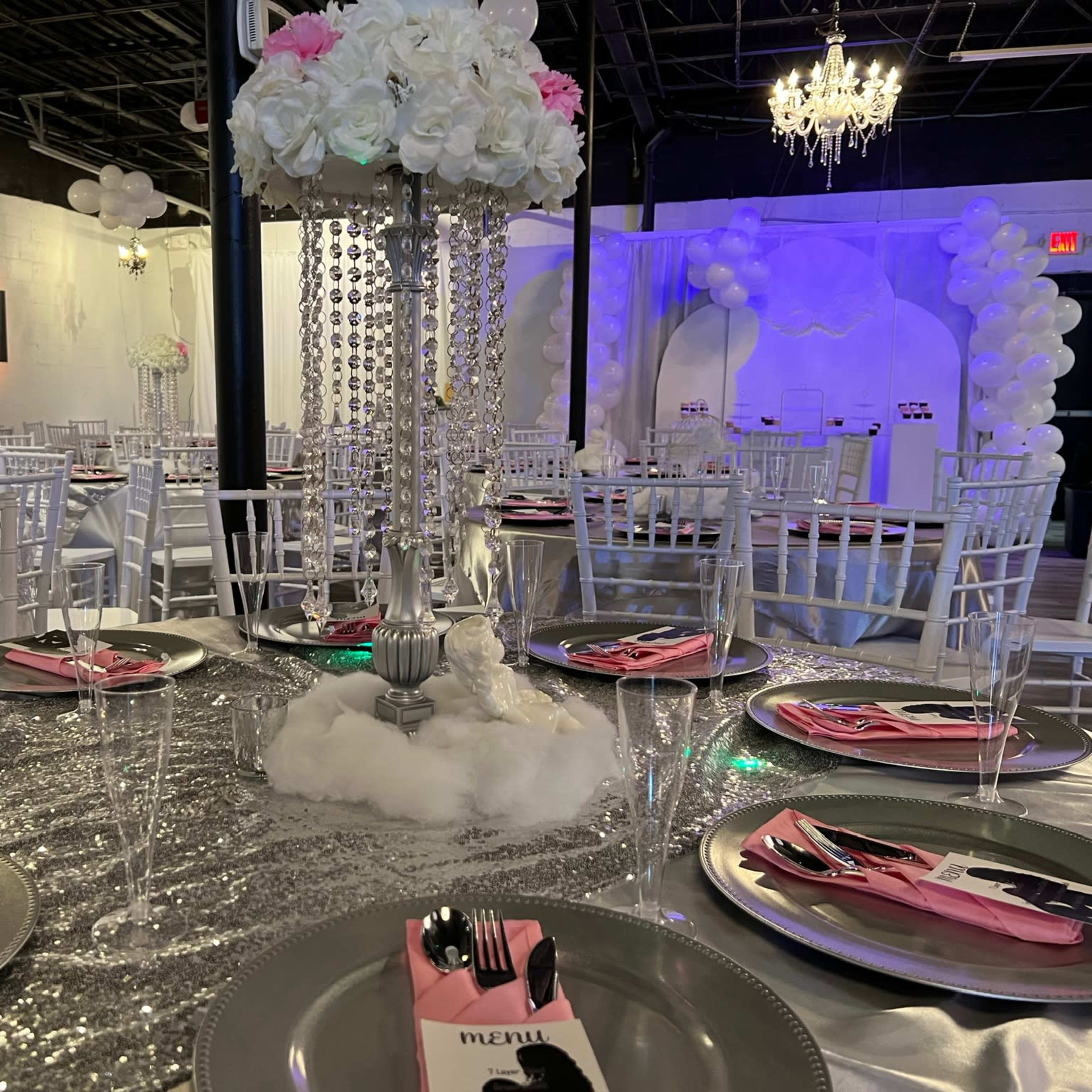 A banquet setup with silver tablecloths, white chairs, and lavish centerpieces featuring flowers and chandeliers in a decorated venue.