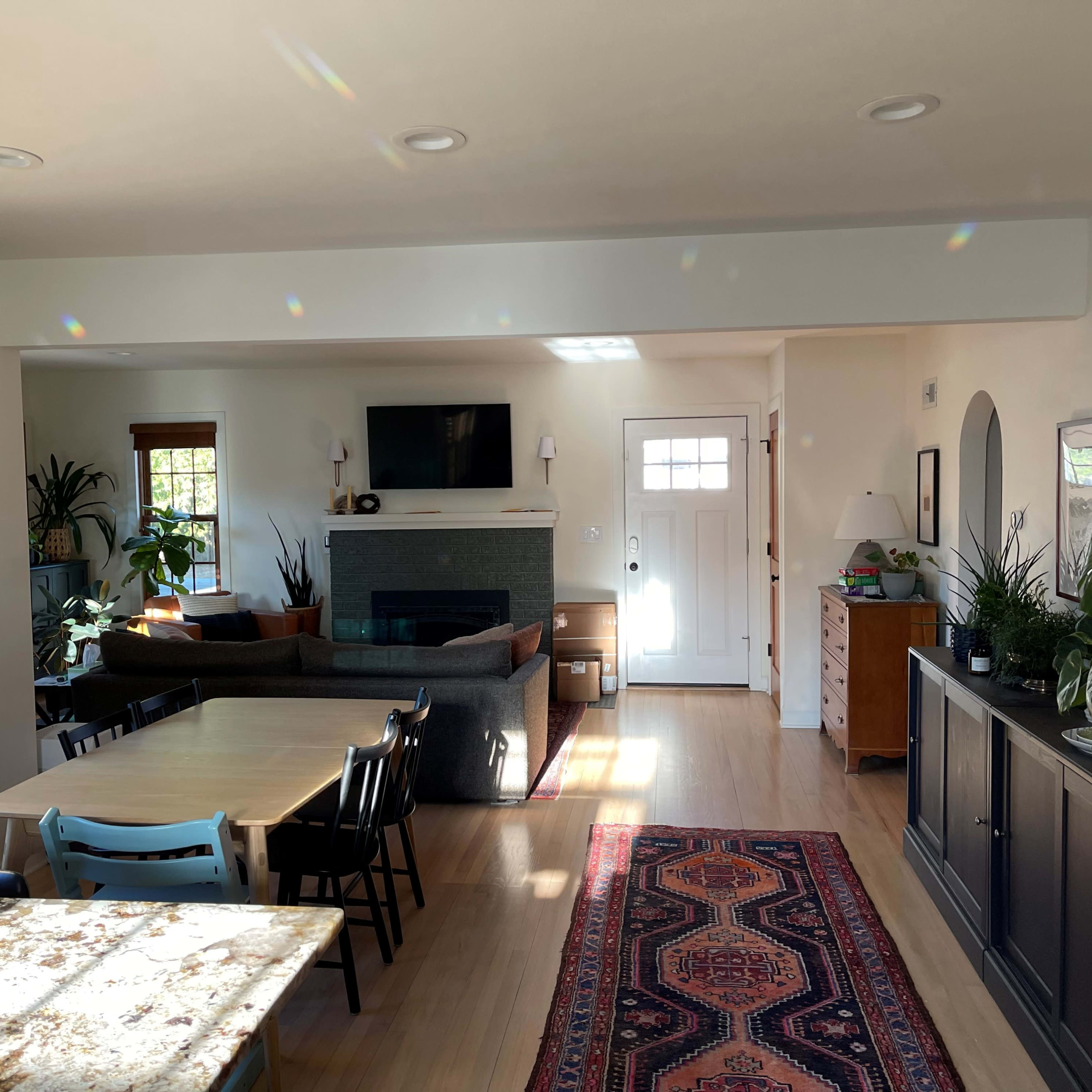 A spacious living area features a fireplace, a large window, a dining table, and various plants, with a front door visible in the background.