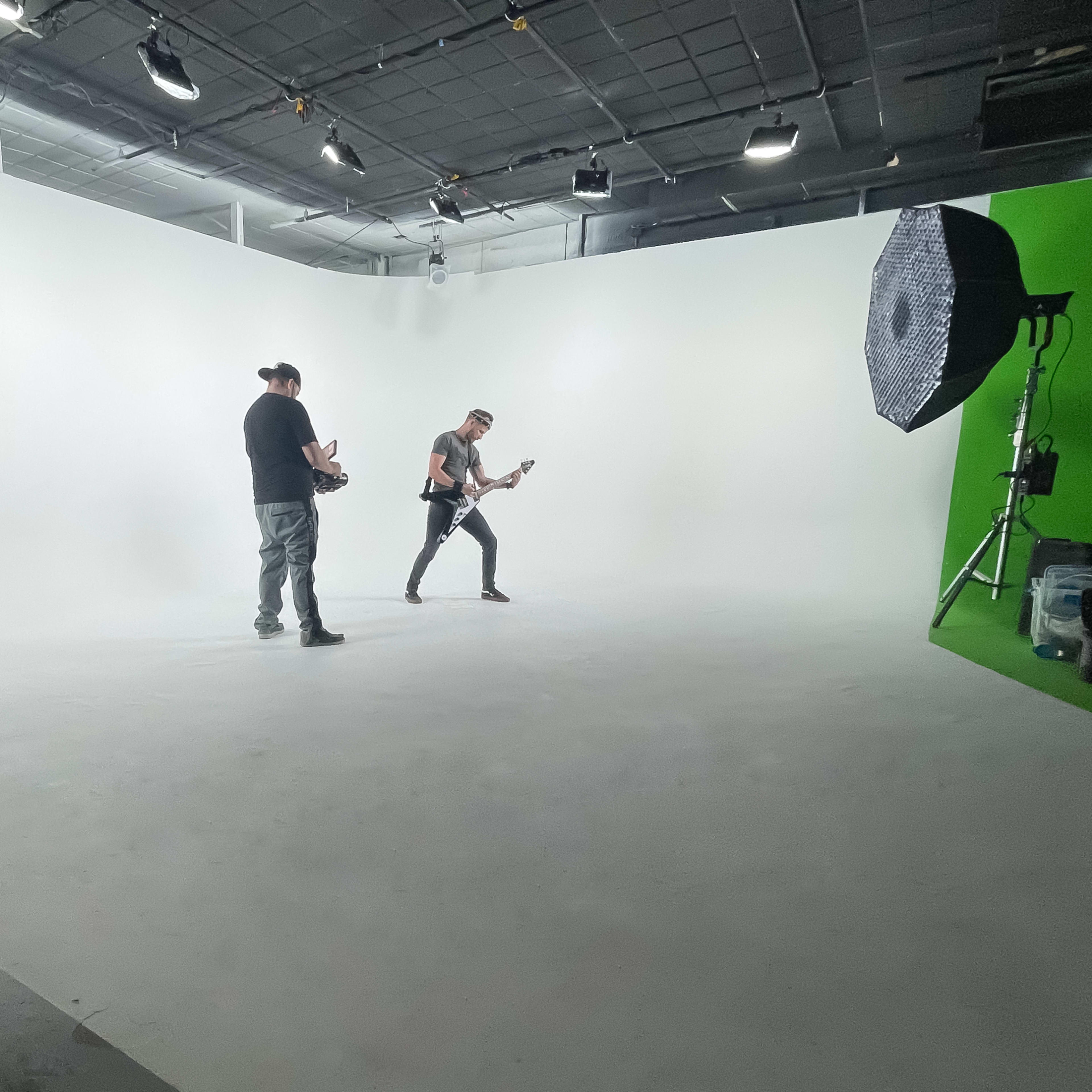 A musician is playing guitar on a white backdrop while another person stands nearby in a performance studio.