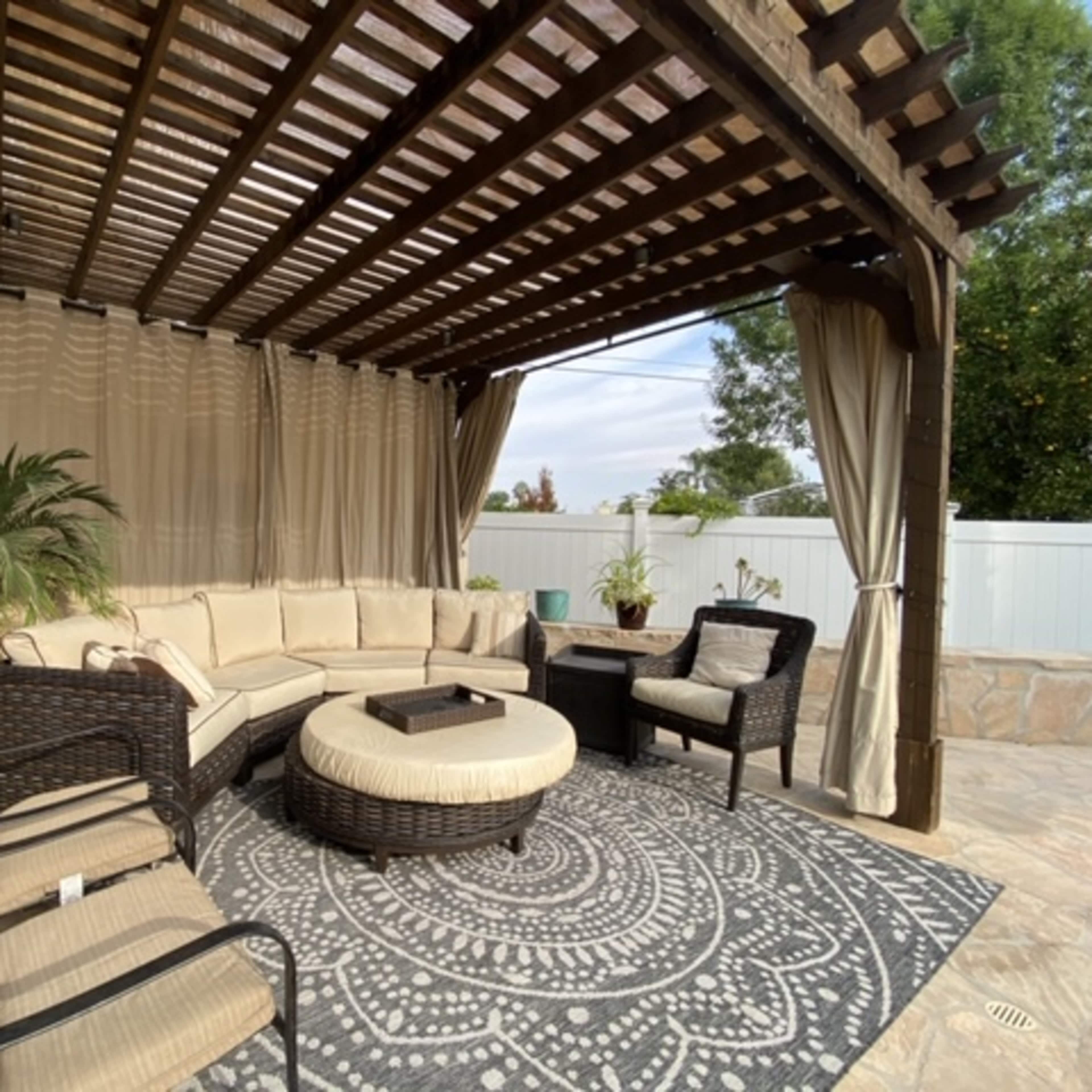 A covered outdoor seating area features cushioned furniture arranged around a central ottoman on a patterned rug.