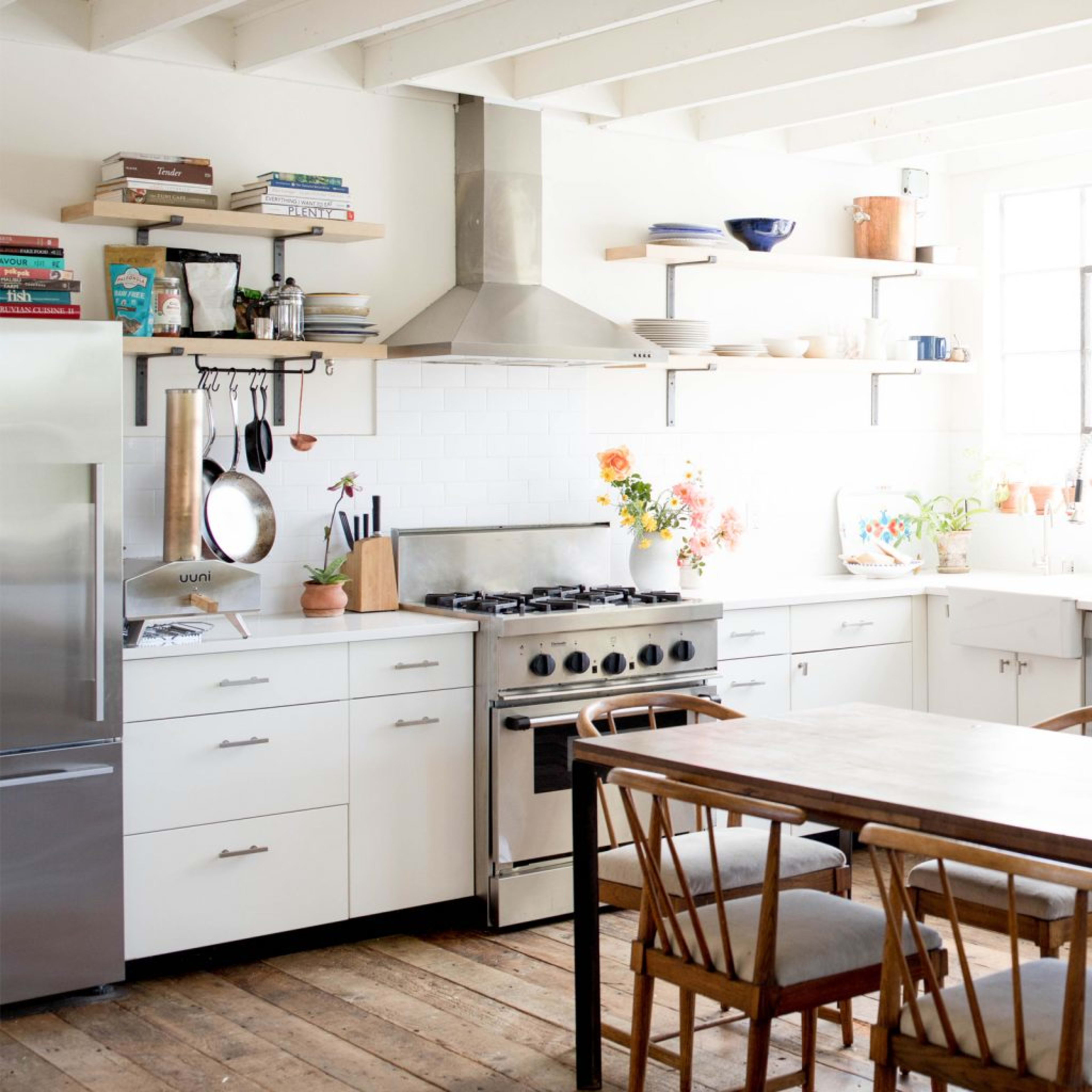 A bright, open kitchen featuring a central wooden dining table, modern appliances, and large windows with plants and decorative items on the shelves.