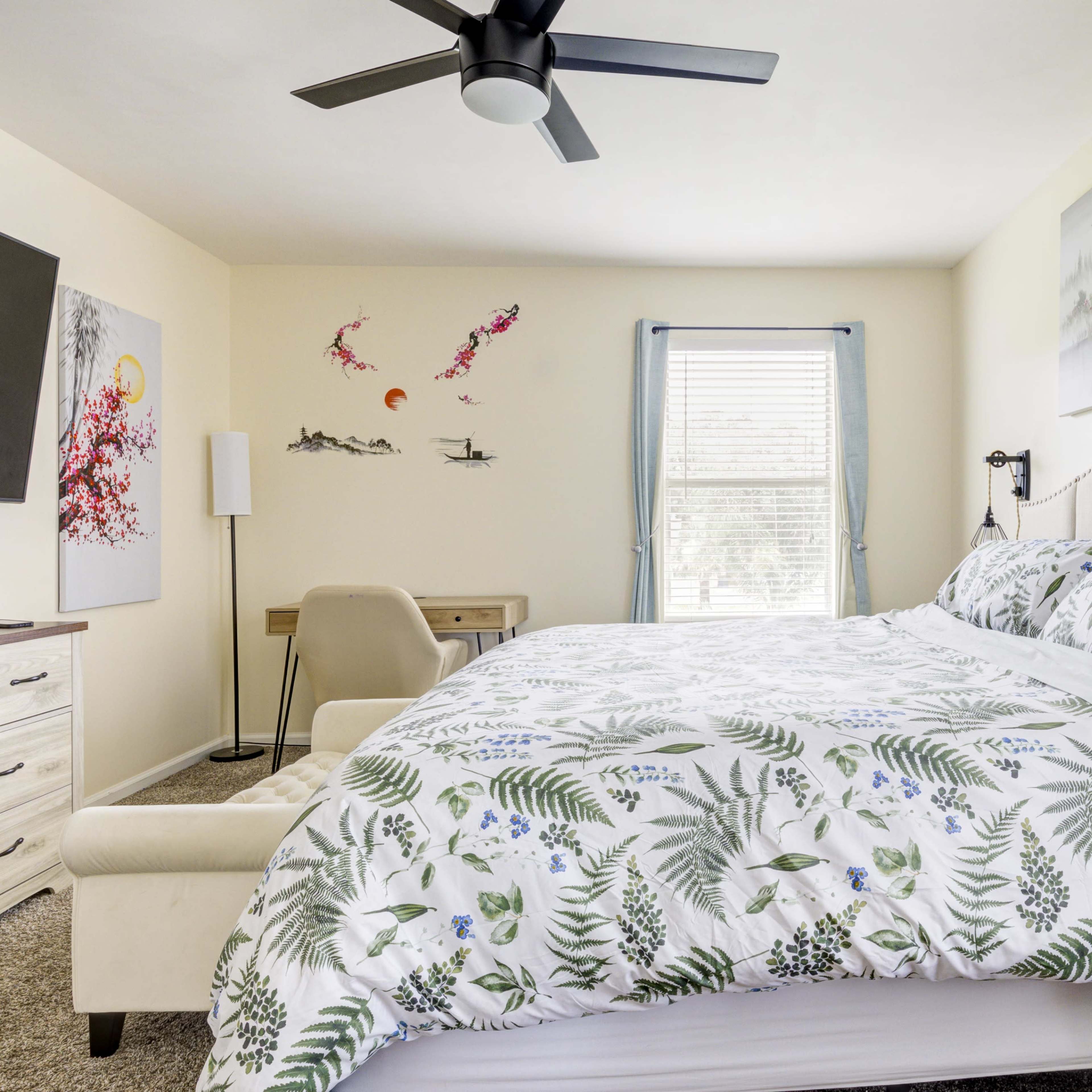 A bedroom features a bed with a floral-patterned comforter, a wall-mounted television, a desk, and artwork depicting trees and birds.