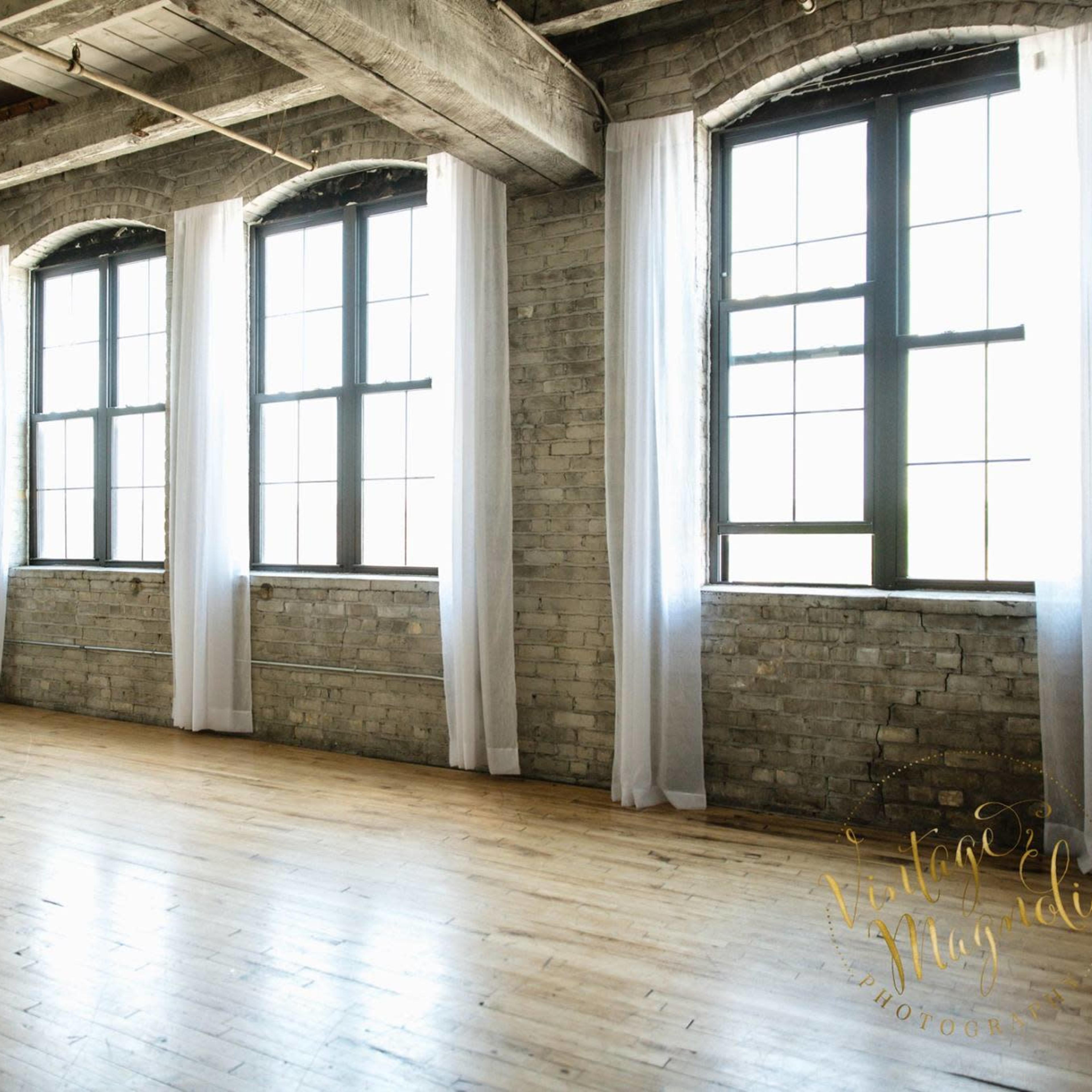 A spacious room with large arched windows, wooden flooring, and exposed brick walls.
