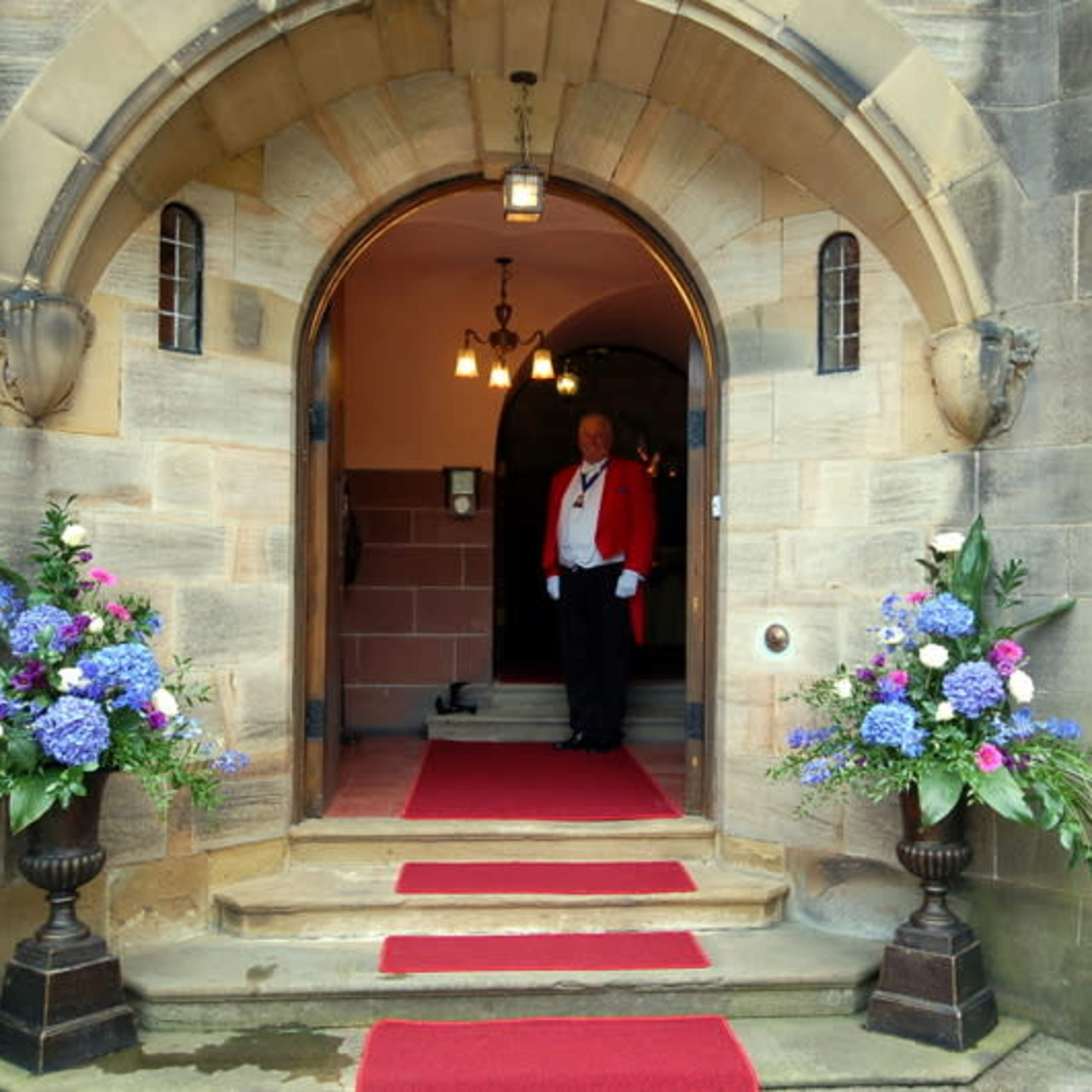 A person in formal attire stands at the entrance of a stone archway, flanked by large floral arrangements and a red carpet leading inside.