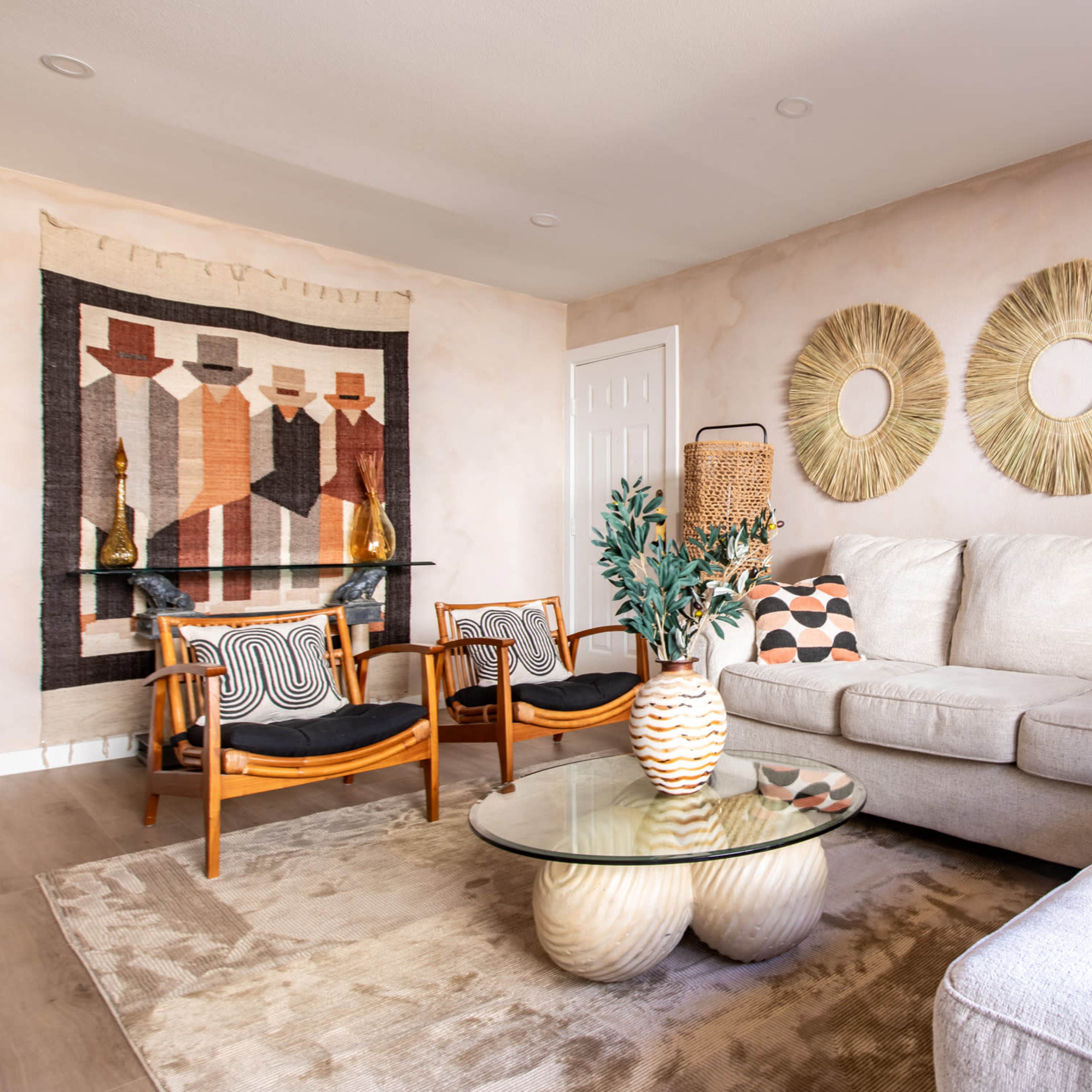 The living room features a neutral color palette with a sectional sofa, a round glass coffee table, two wooden chairs, and decorative wall hangings.