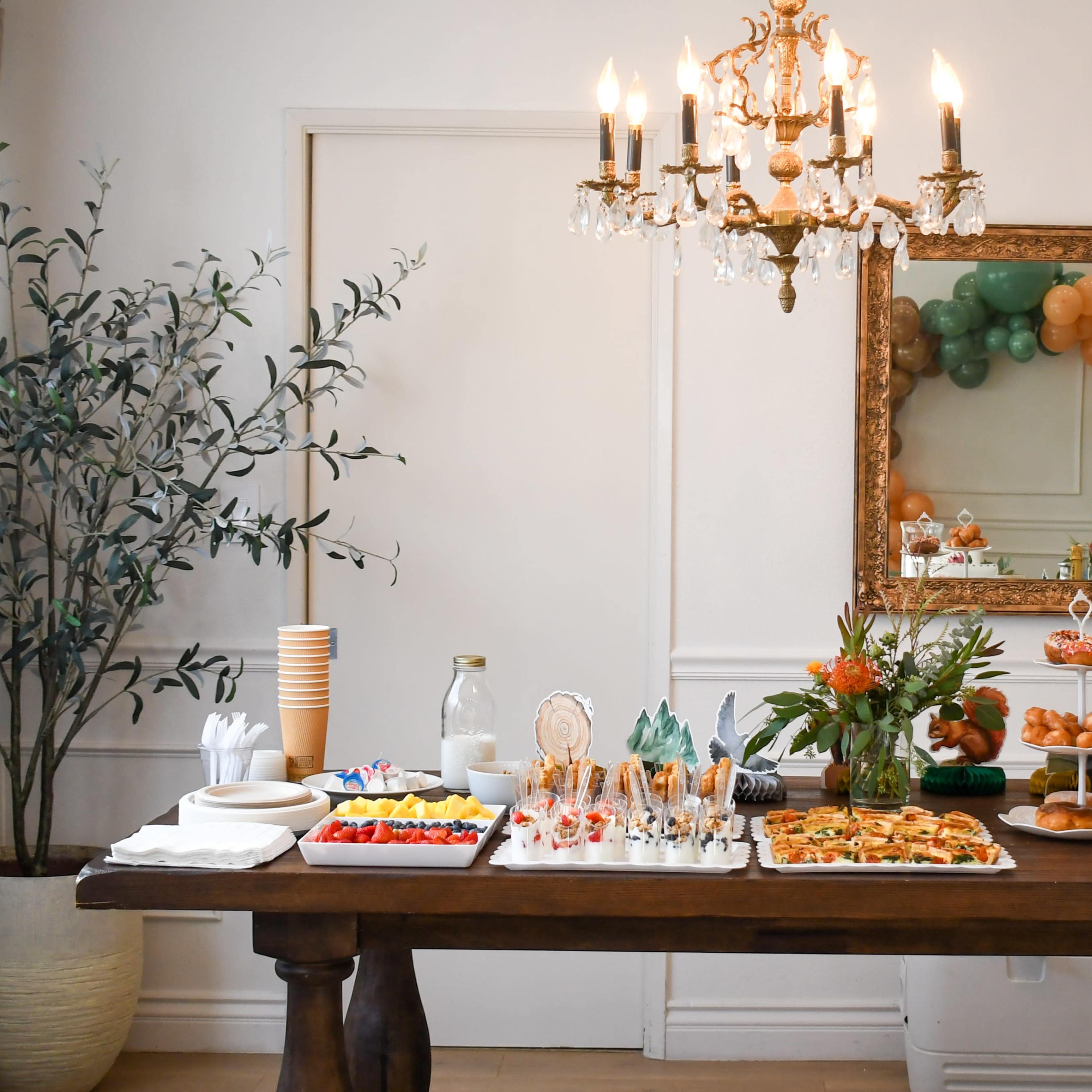 A decorated table is set with an assortment of food and drinks, featuring various appetizers, desserts, and a large chandelier overhead.