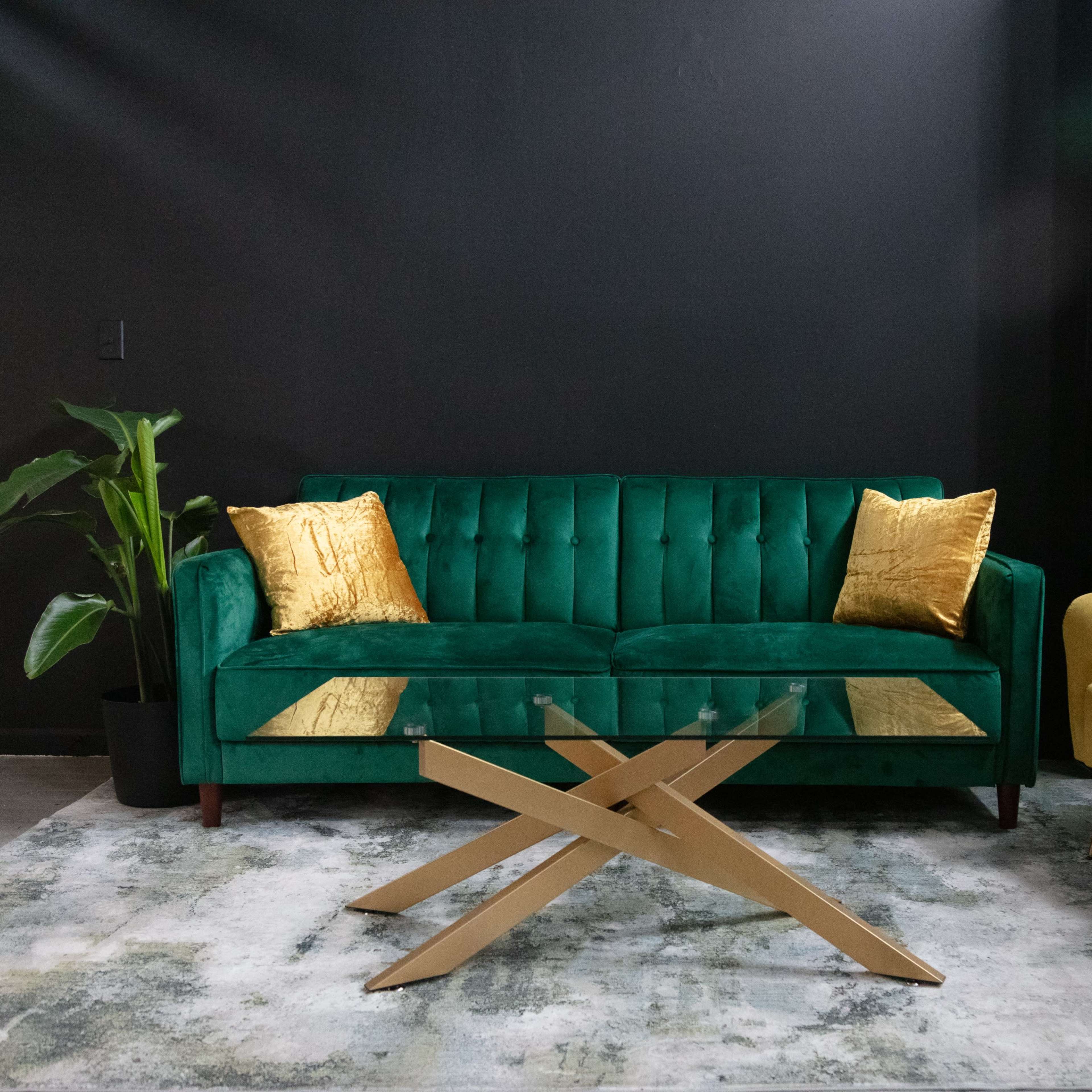 A green velvet sofa with gold cushions and a yellow armchair are positioned around a glass-top coffee table on a textured rug in a dark room.