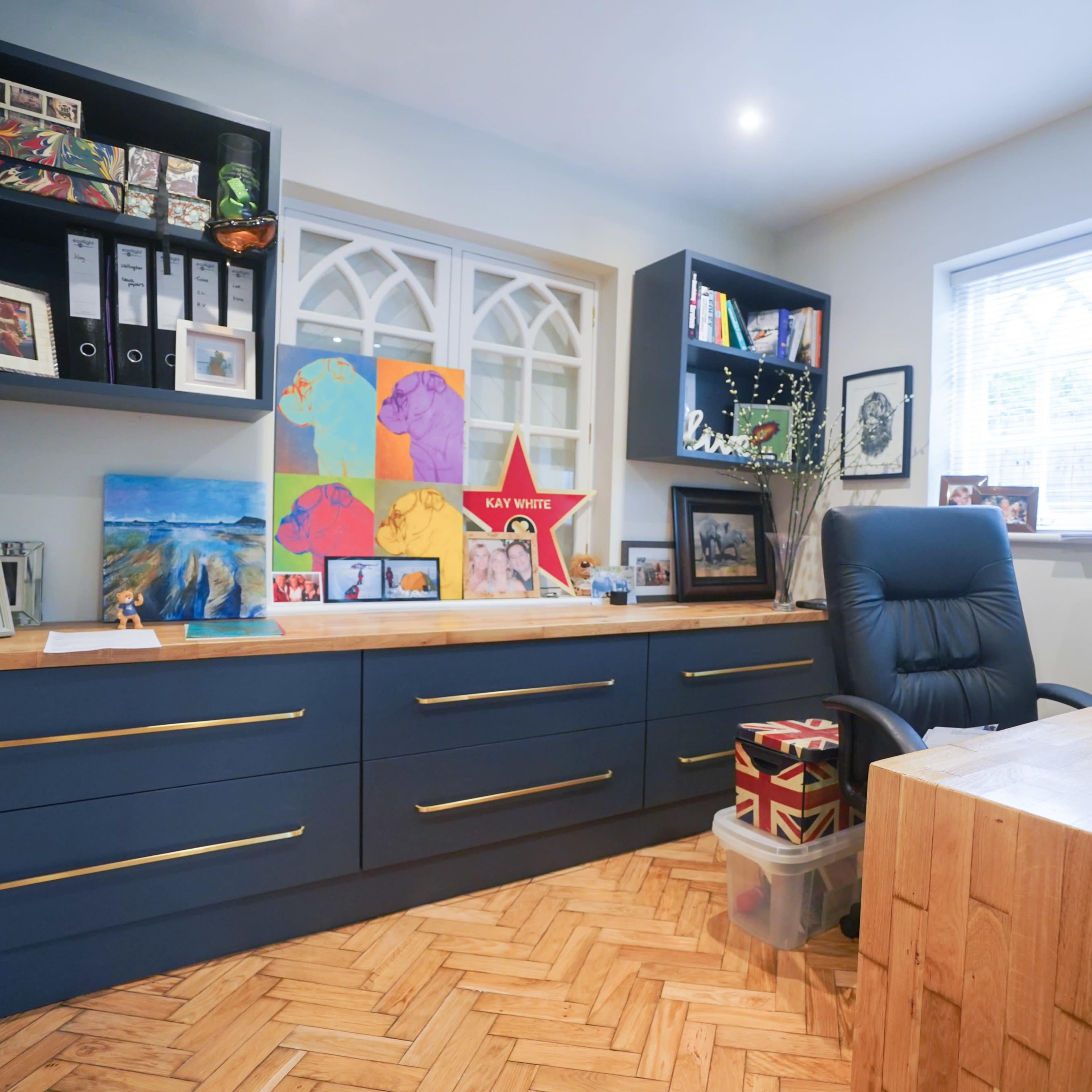 A well-organized home office featuring a dark blue desk with drawers, shelving with framed pictures and colorful artwork, and a black ergonomic chair next to a wooden desk.