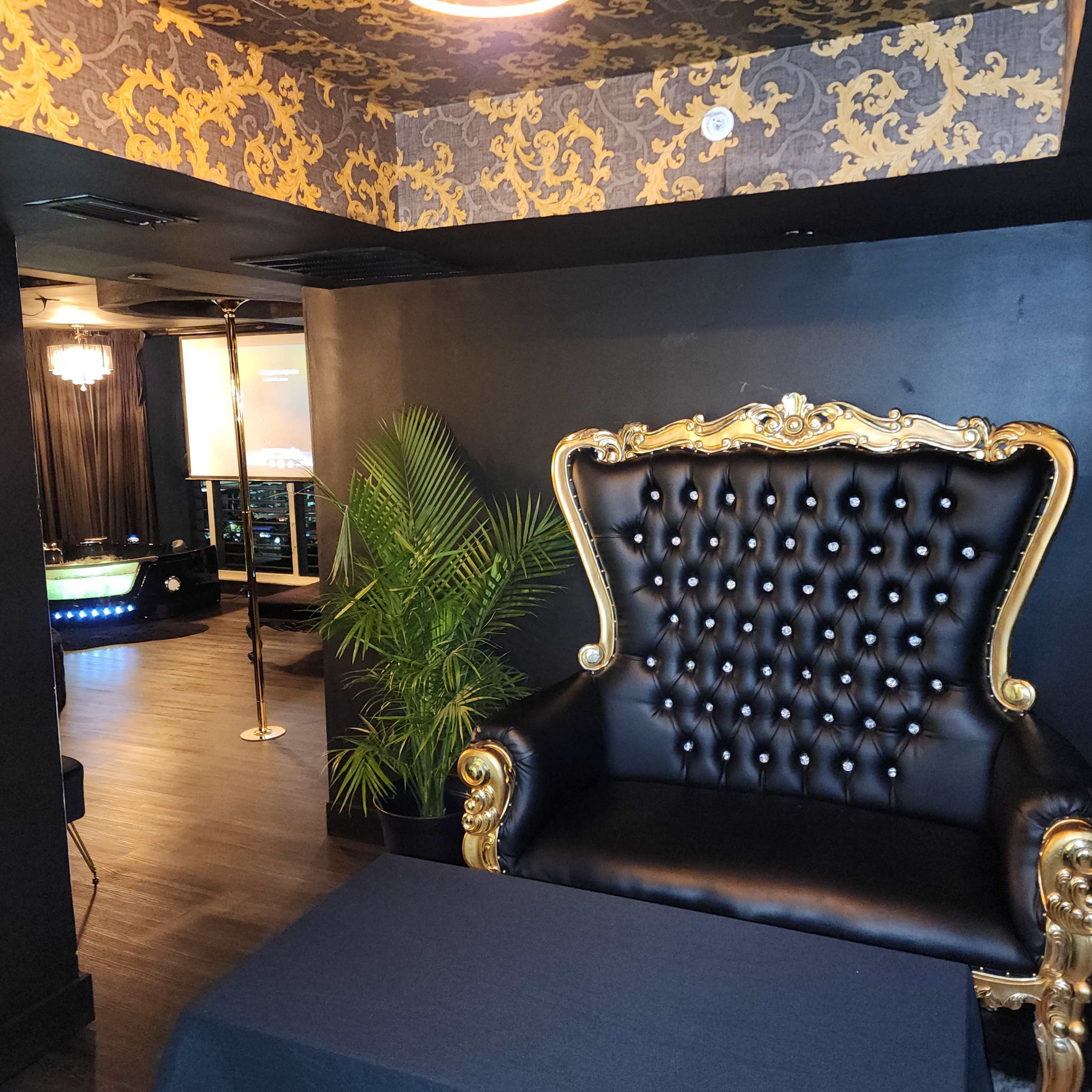 A lavish black and gold decorated room featuring an ornate golden throne chair in the foreground, with a table and a green plant beside it, and a projector screen visible in the background.