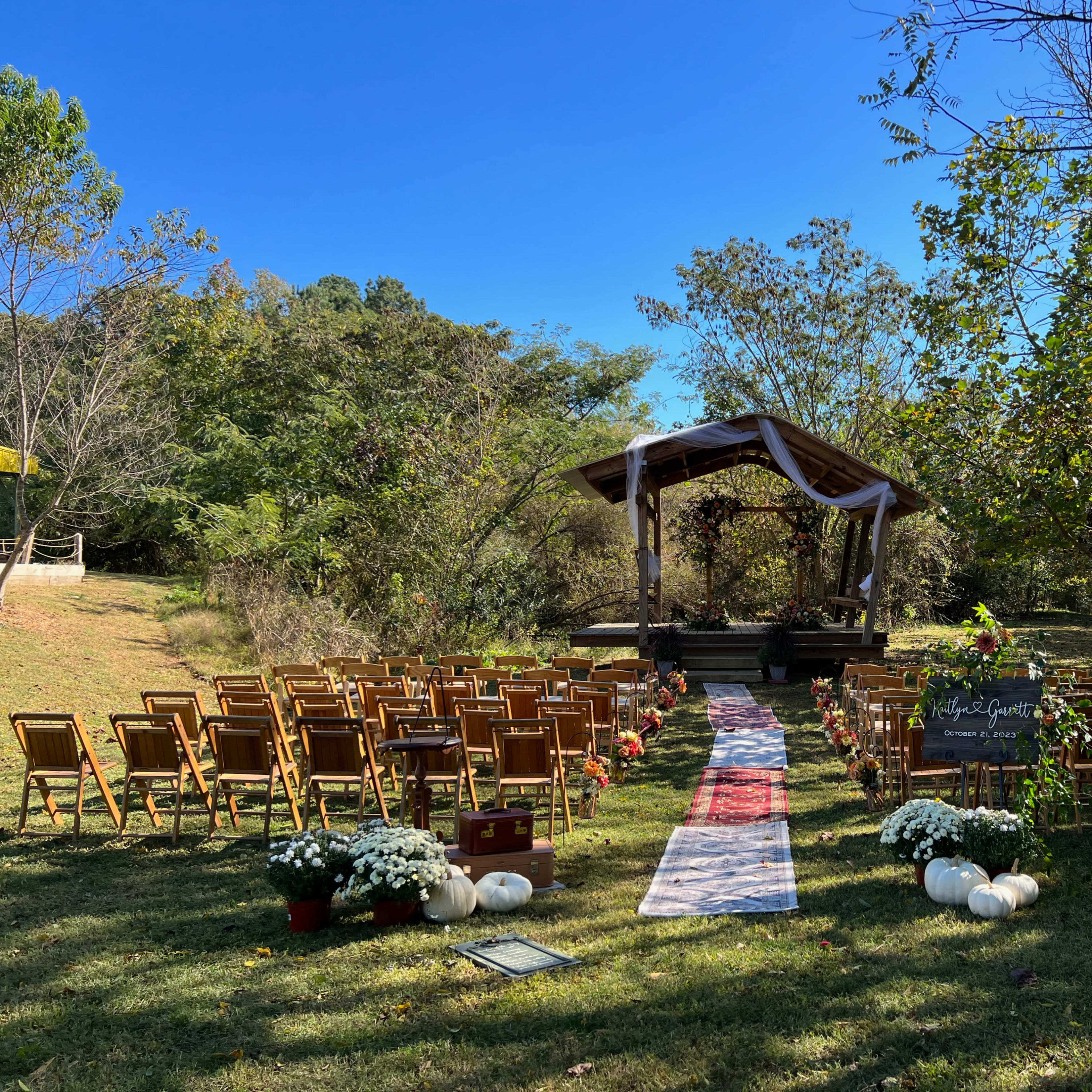 A wedding ceremony setup features rows of wooden chairs arranged on a grassy area leading to a decorated altar under a gazebo, surrounded by trees.