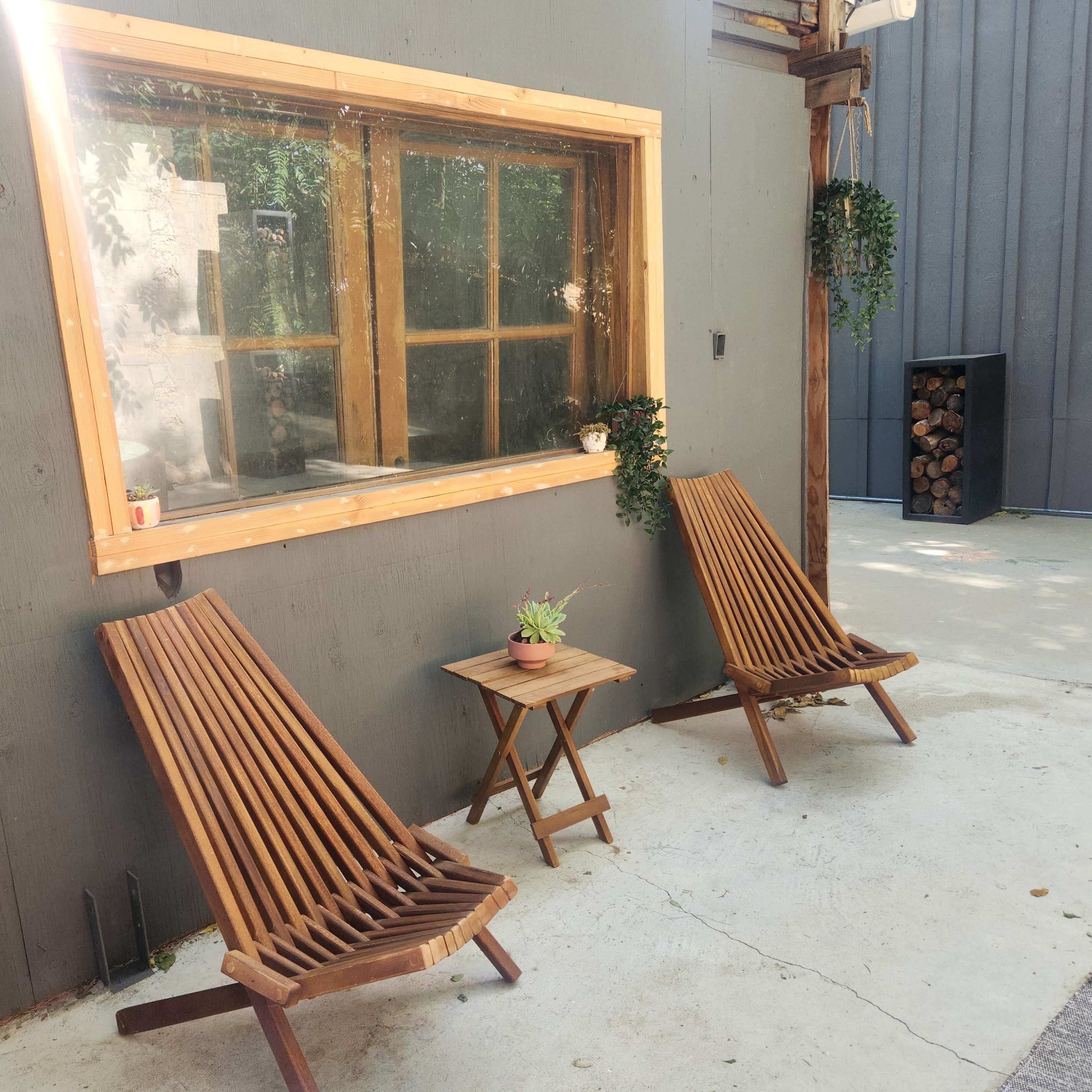 Two wooden lounge chairs are positioned next to a small table with a potted plant, beside a large window in a gray wall.