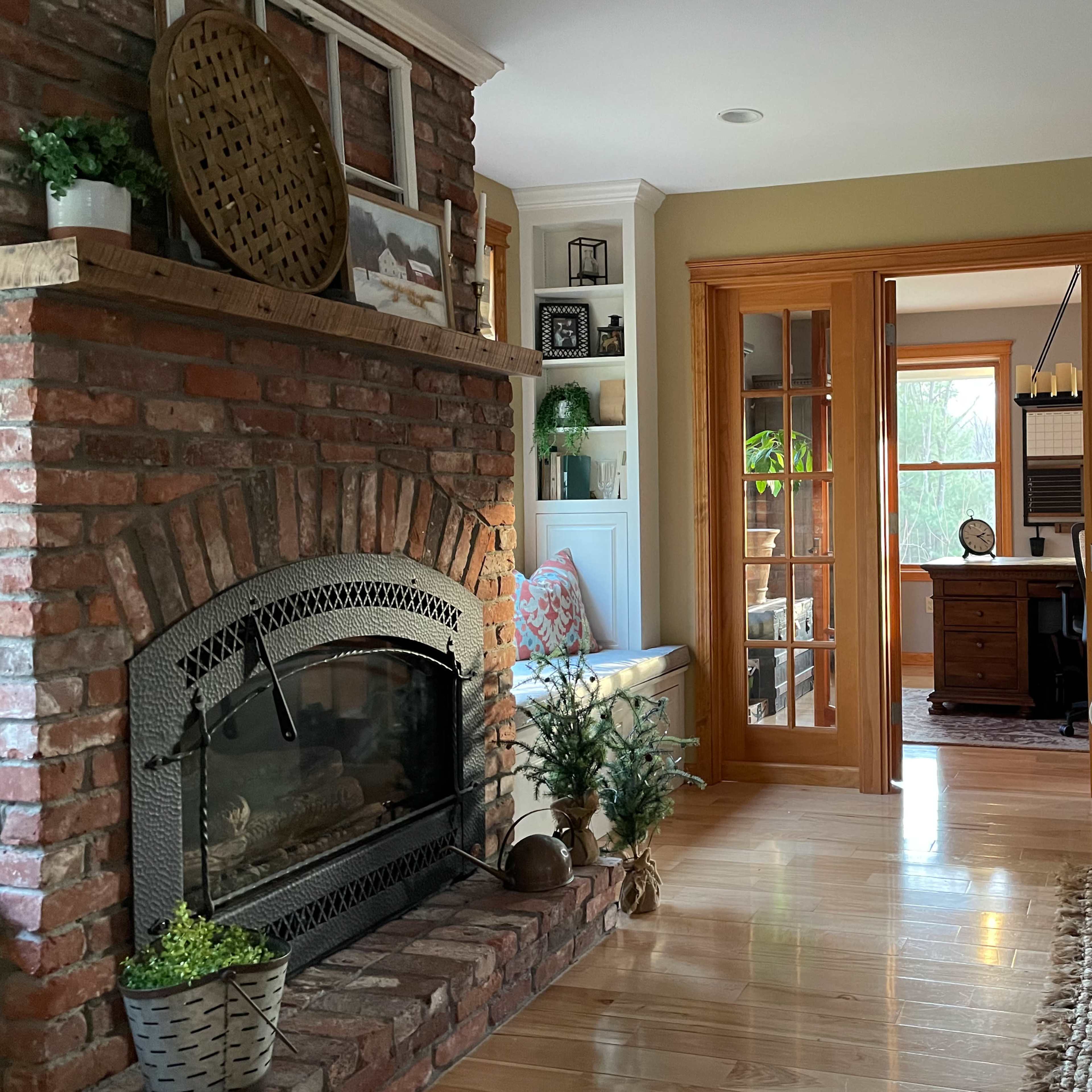 A cozy living room featuring a brick fireplace, a comfortable sofa, and an adjoining room with a desk and large windows.