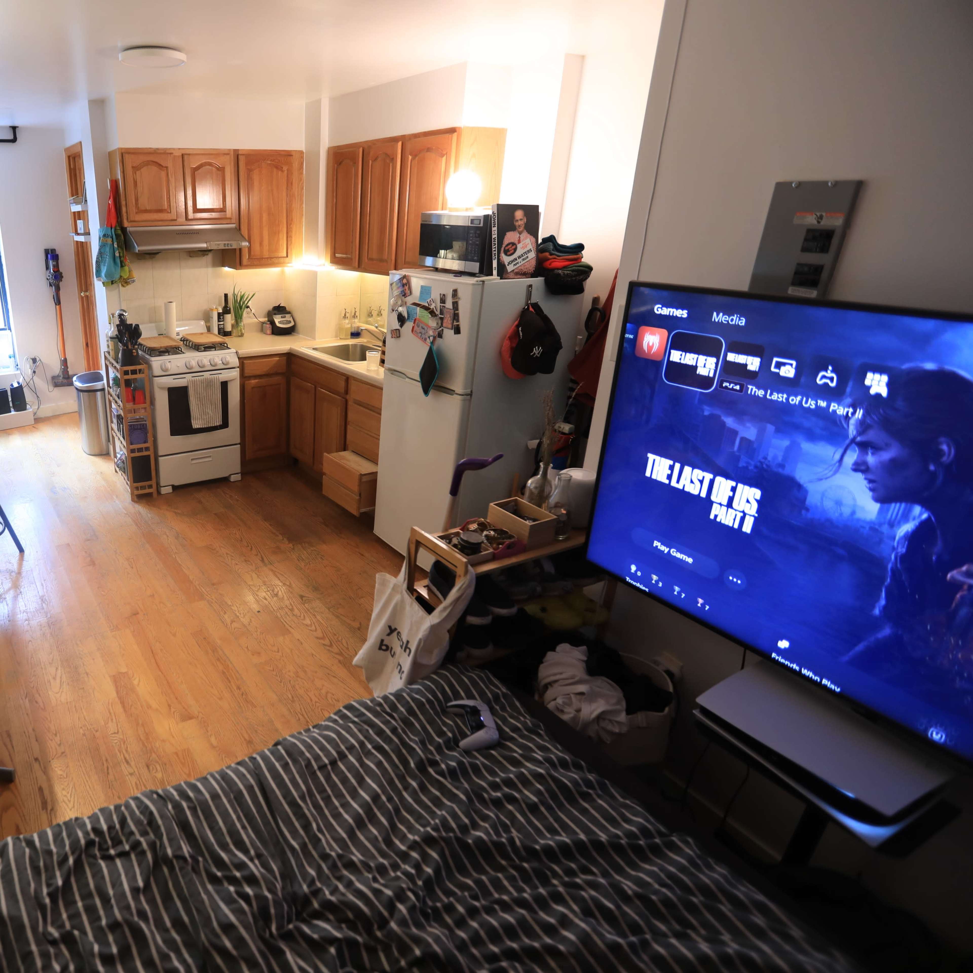 A studio apartment features a kitchen area with wooden cabinets, a living space with a large television, and a bed positioned near a window.