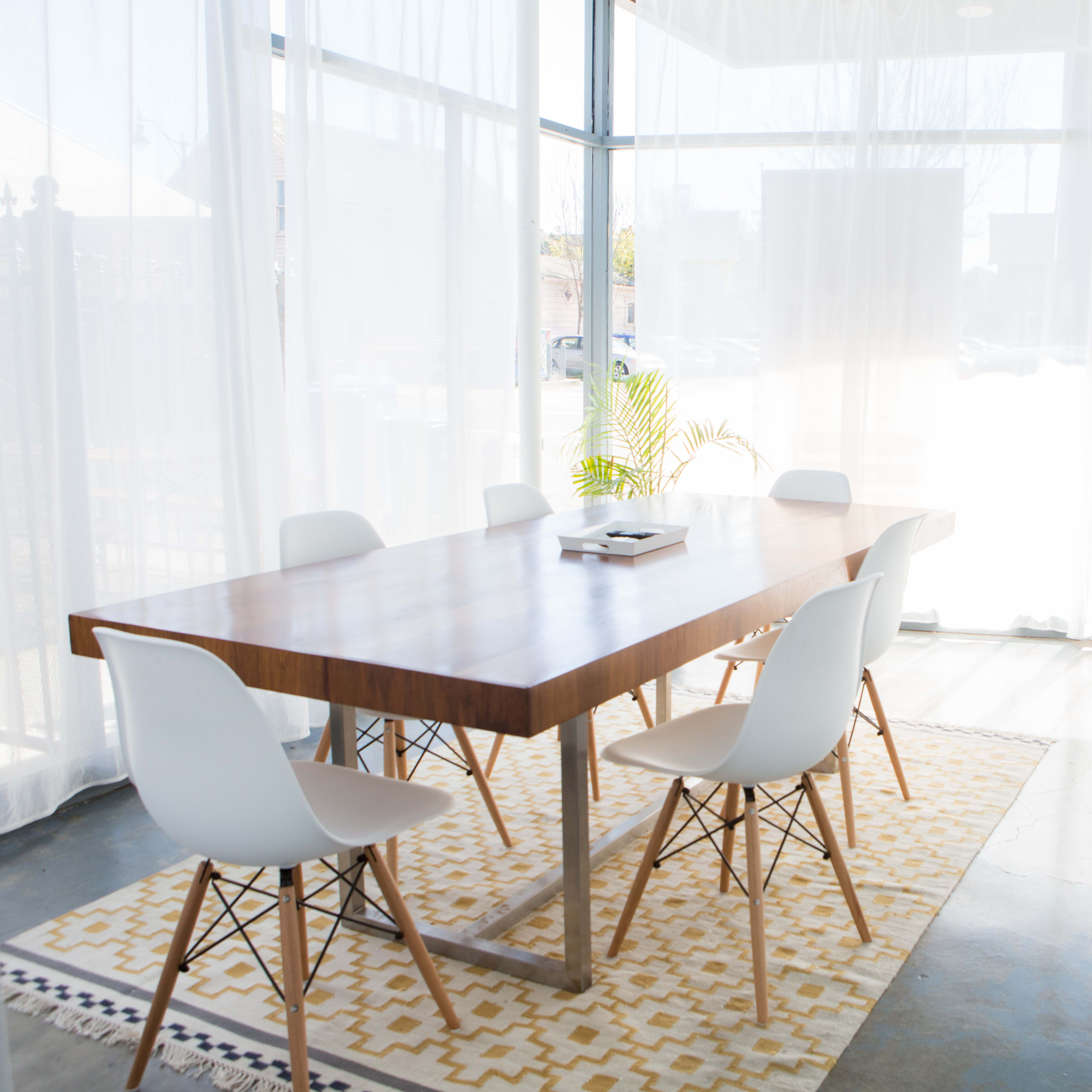A modern conference room features a wooden table surrounded by white chairs, with large windows partially covered by sheer curtains.